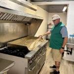 Man in white cap and green polo standing in a commercial kitchen next to a large flat-top griddle.
