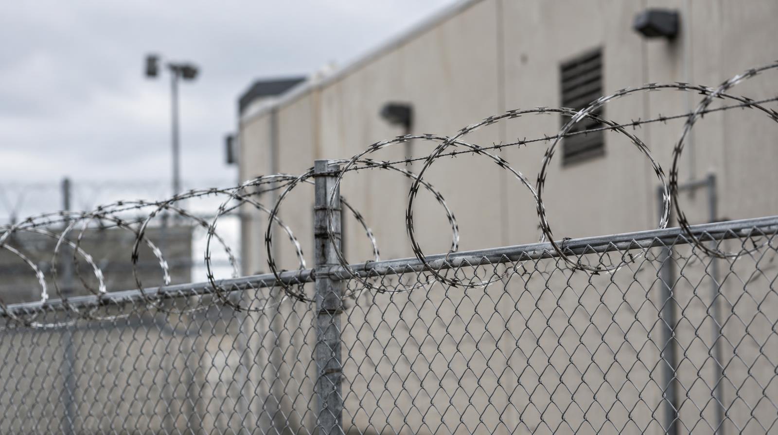A chain-link fence with razor wire on top in front of a concrete wall.
