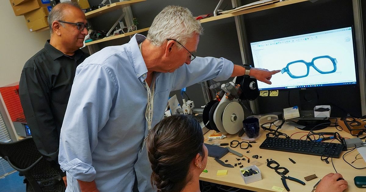 Three people gathered around a workstation examining a 3D eyeglass frame model on a computer monitor.