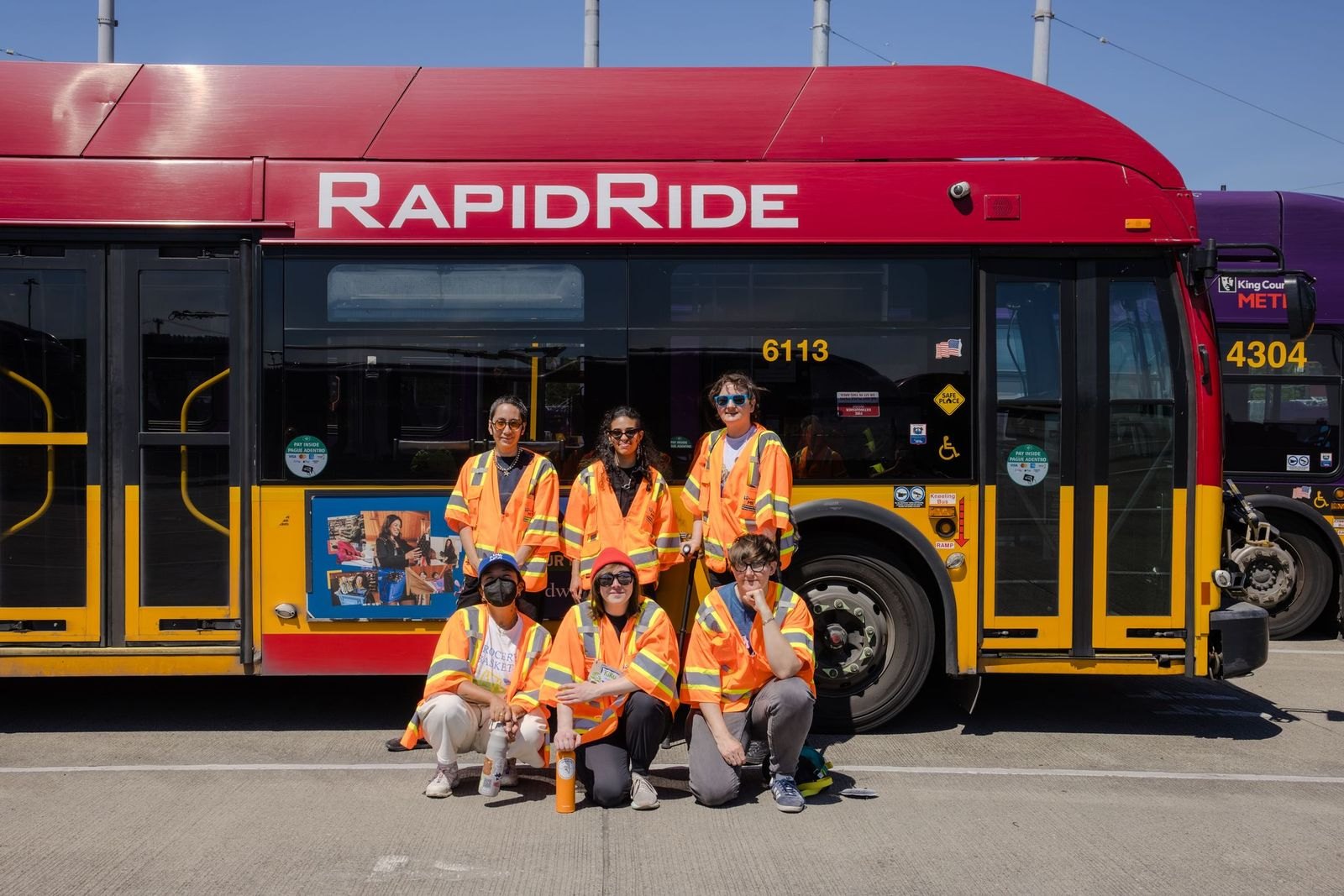 Six people in orange safety vests posed in two rows in front of a red RapidRide bus.