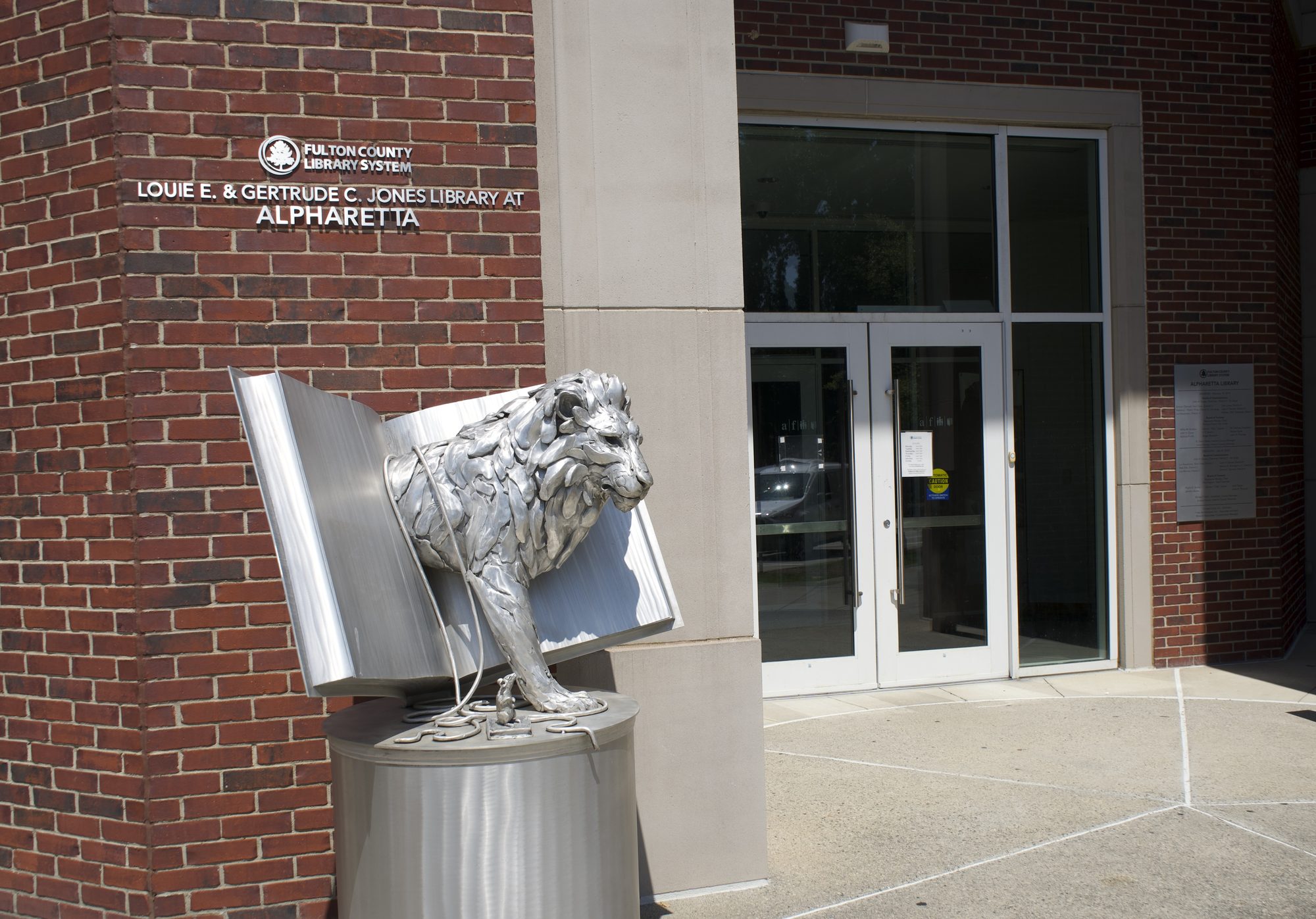 Exterior entrance of a brick public library with a metal lion-and-book sculpture in front.