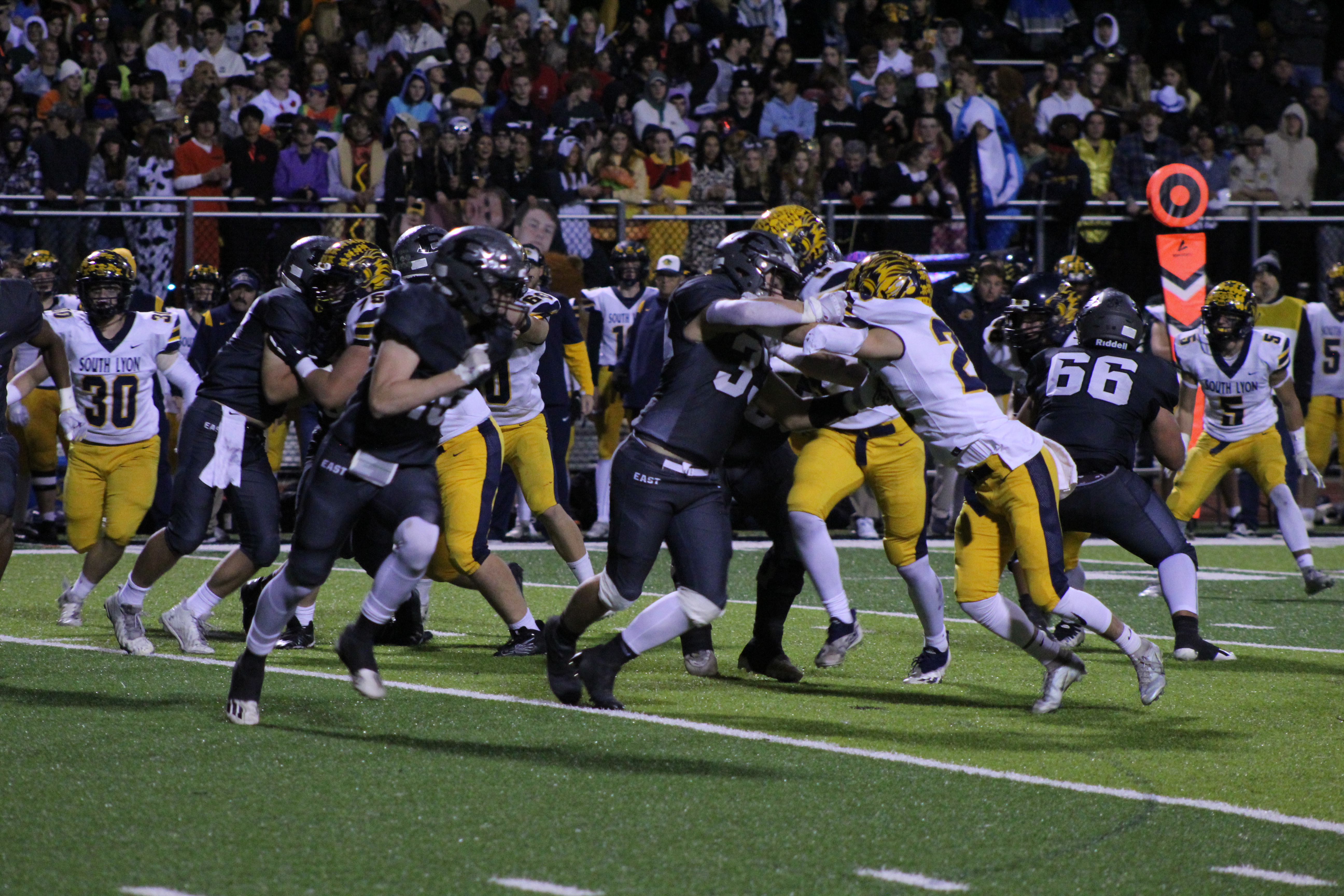 High school football players in dark and gold uniforms face off at the line of scrimmage under stadium lights.