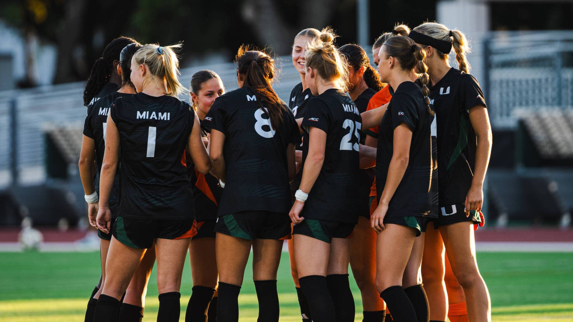 Women soccer players in black Miami jerseys huddling together on a grass field.