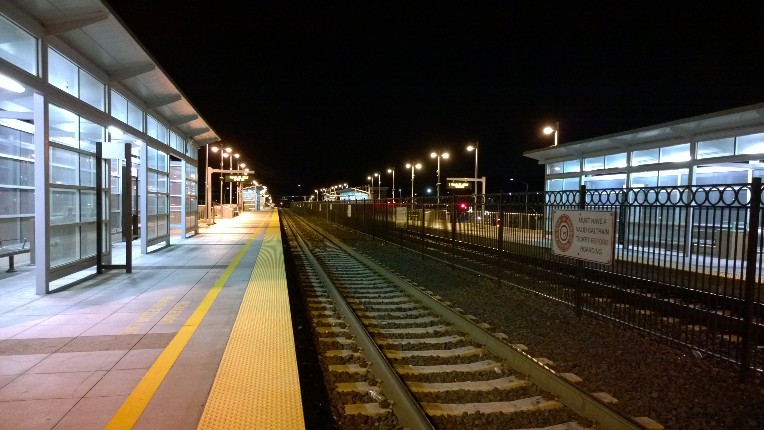 Empty Caltrain station platform at night with glass shelters and yellow tactile edge strips along the tracks.
