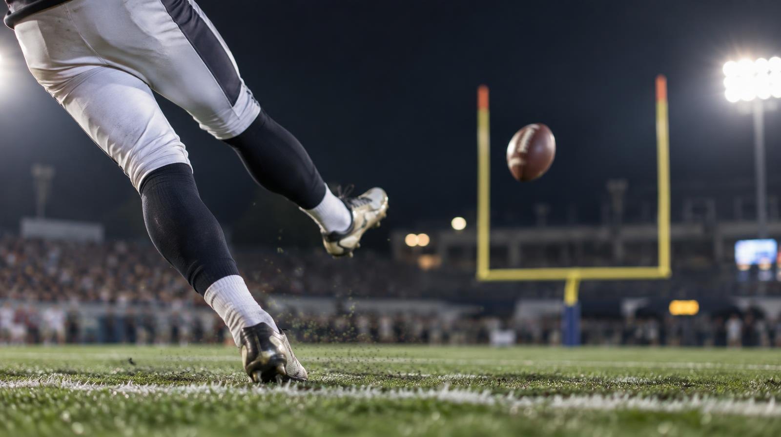 A football kicker's leg following through on a kick with yellow goal posts in the background.