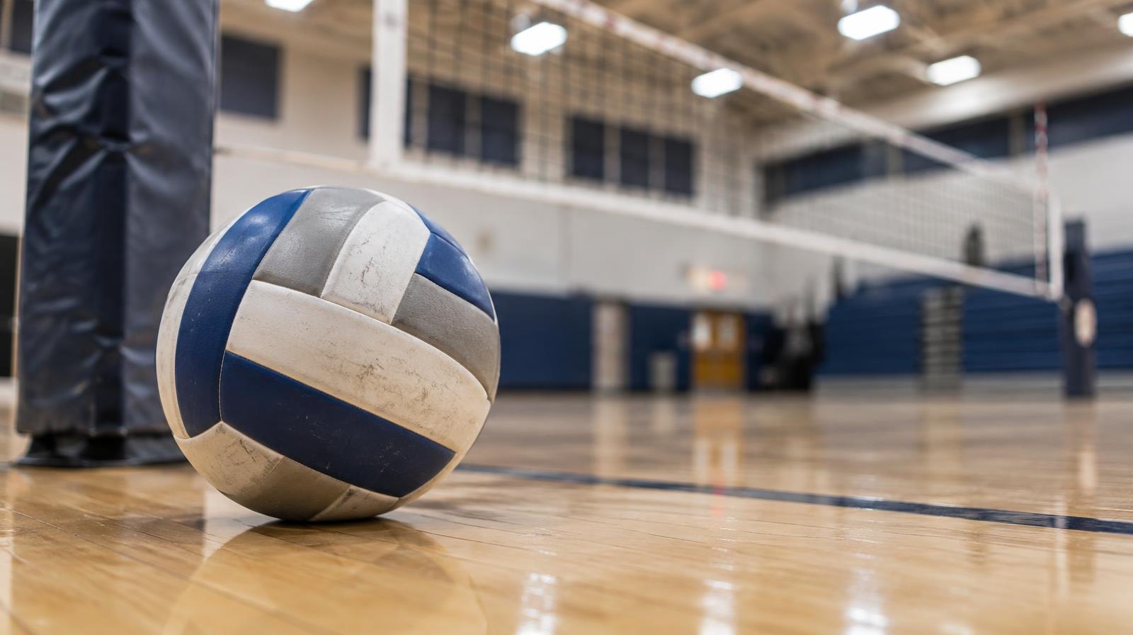 A volleyball resting on a hardwood gymnasium floor near a net post.