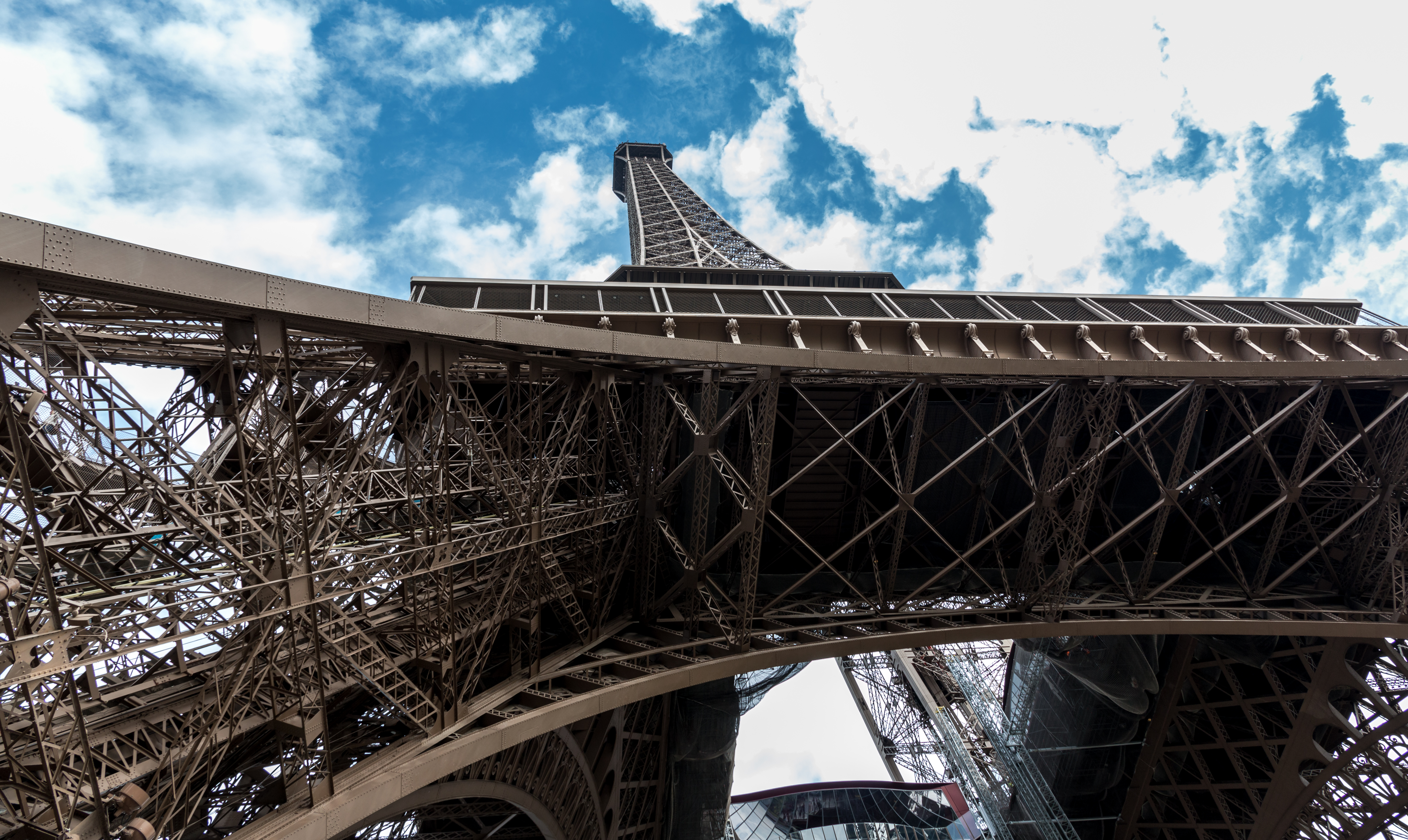 Looking up through the iron lattice of the Eiffel Tower toward a partly cloudy sky.