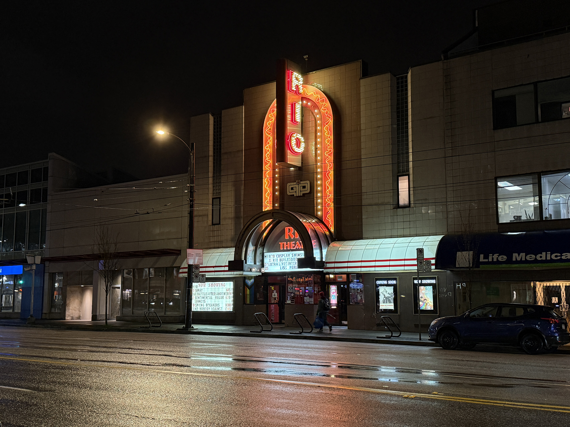 Historic Art Deco theater exterior at night with glowing red neon sign and wet street reflection.