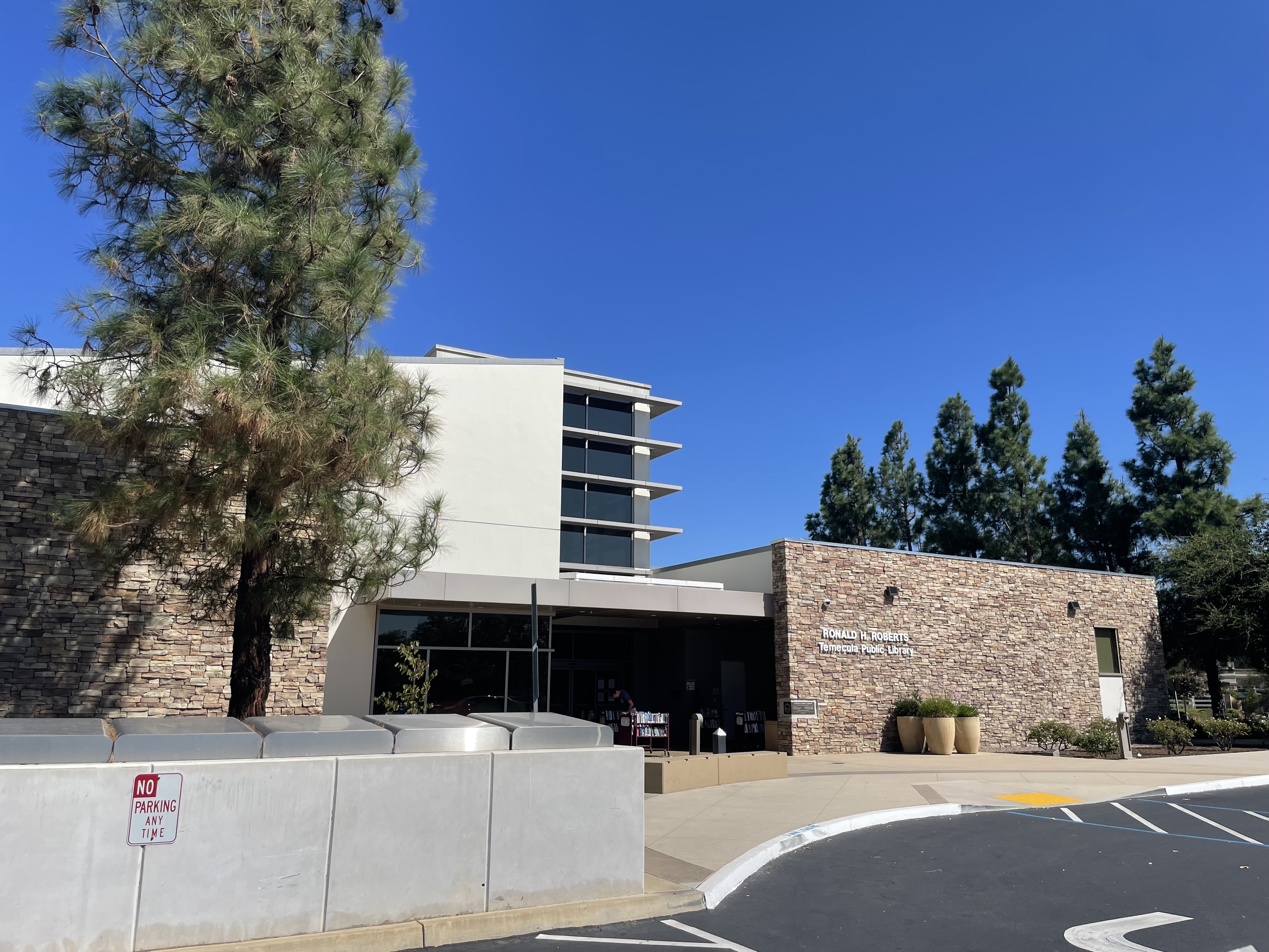 Modern public library building with stone facade, large windows, and tall trees.