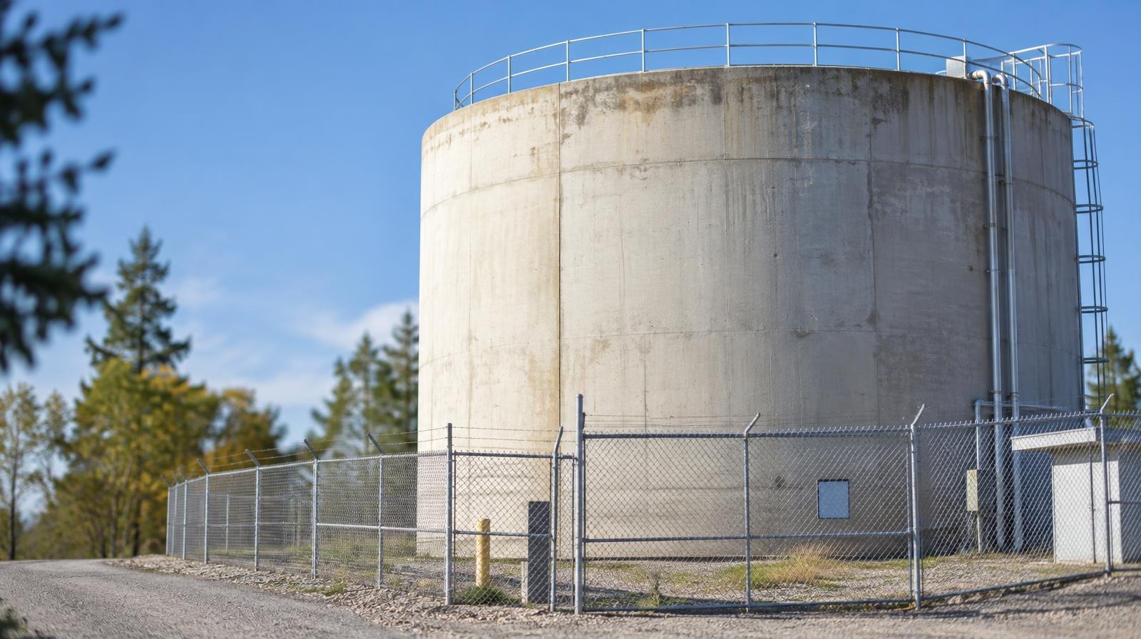 Large cylindrical concrete water storage tank behind a chain-link fence under blue sky.