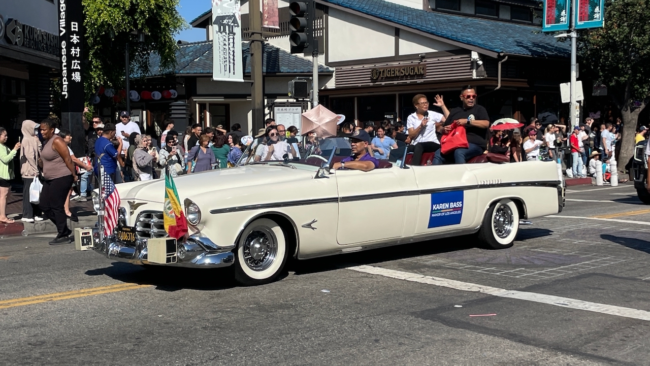 White vintage convertible with a Karen Bass placard moving through a parade route lined with spectators.