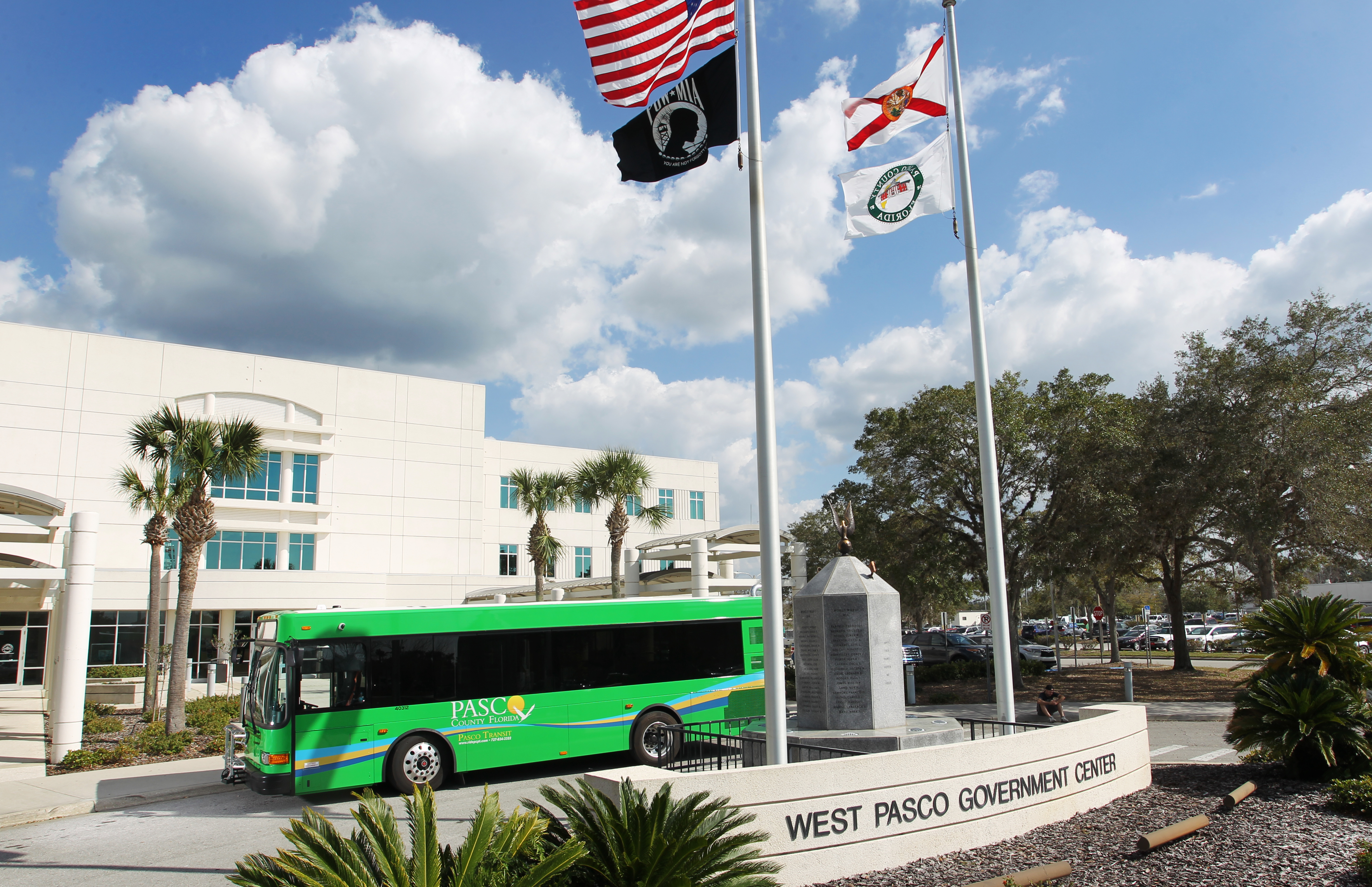 Exterior of a government building with flags on a pole and a green county bus passing in front.