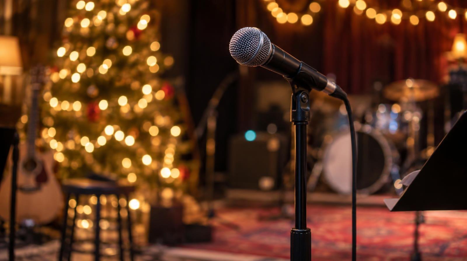 A microphone stand on a darkened concert stage with glowing holiday string lights behind it.