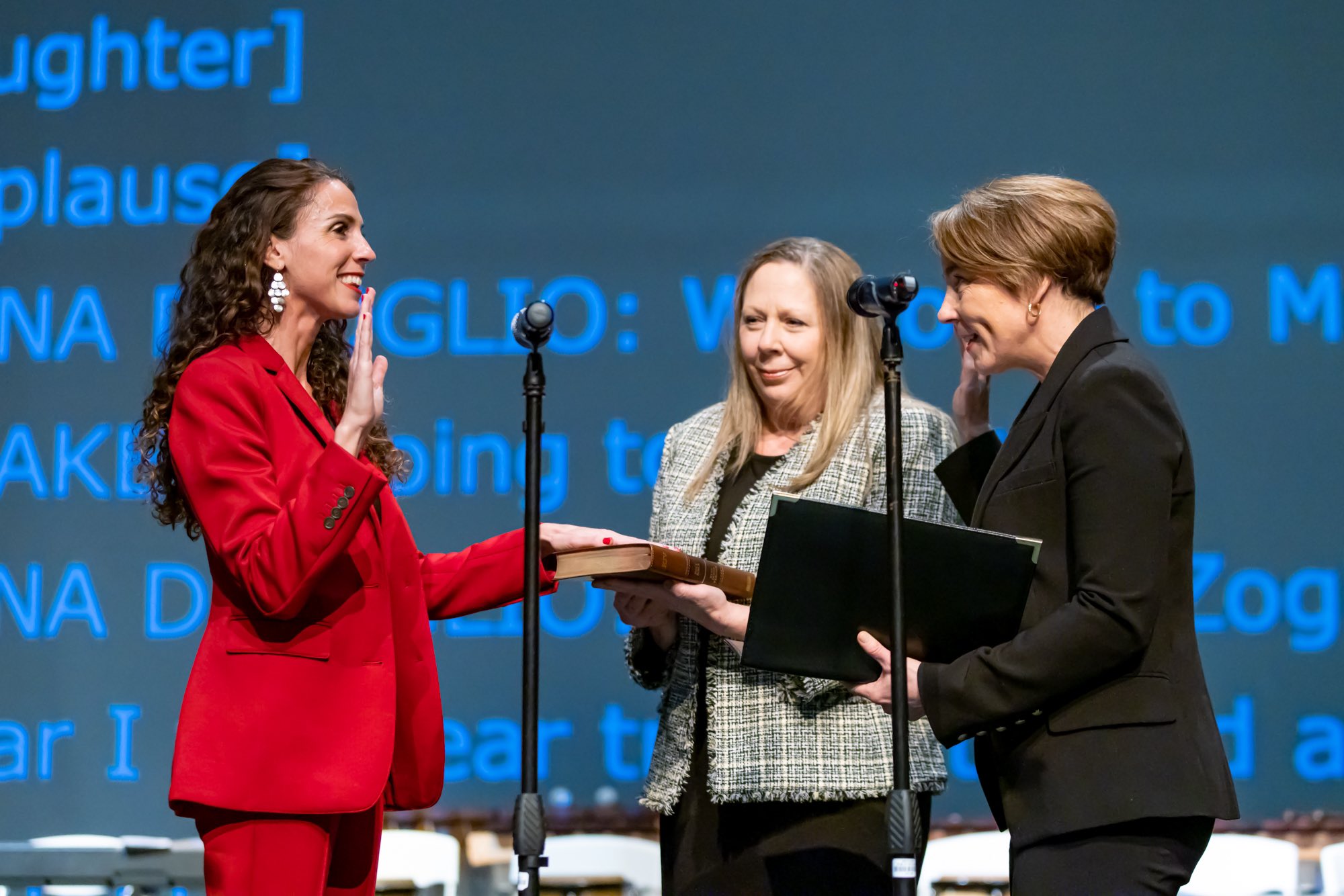 A woman in a red suit raises her right hand during a swearing-in ceremony on stage.