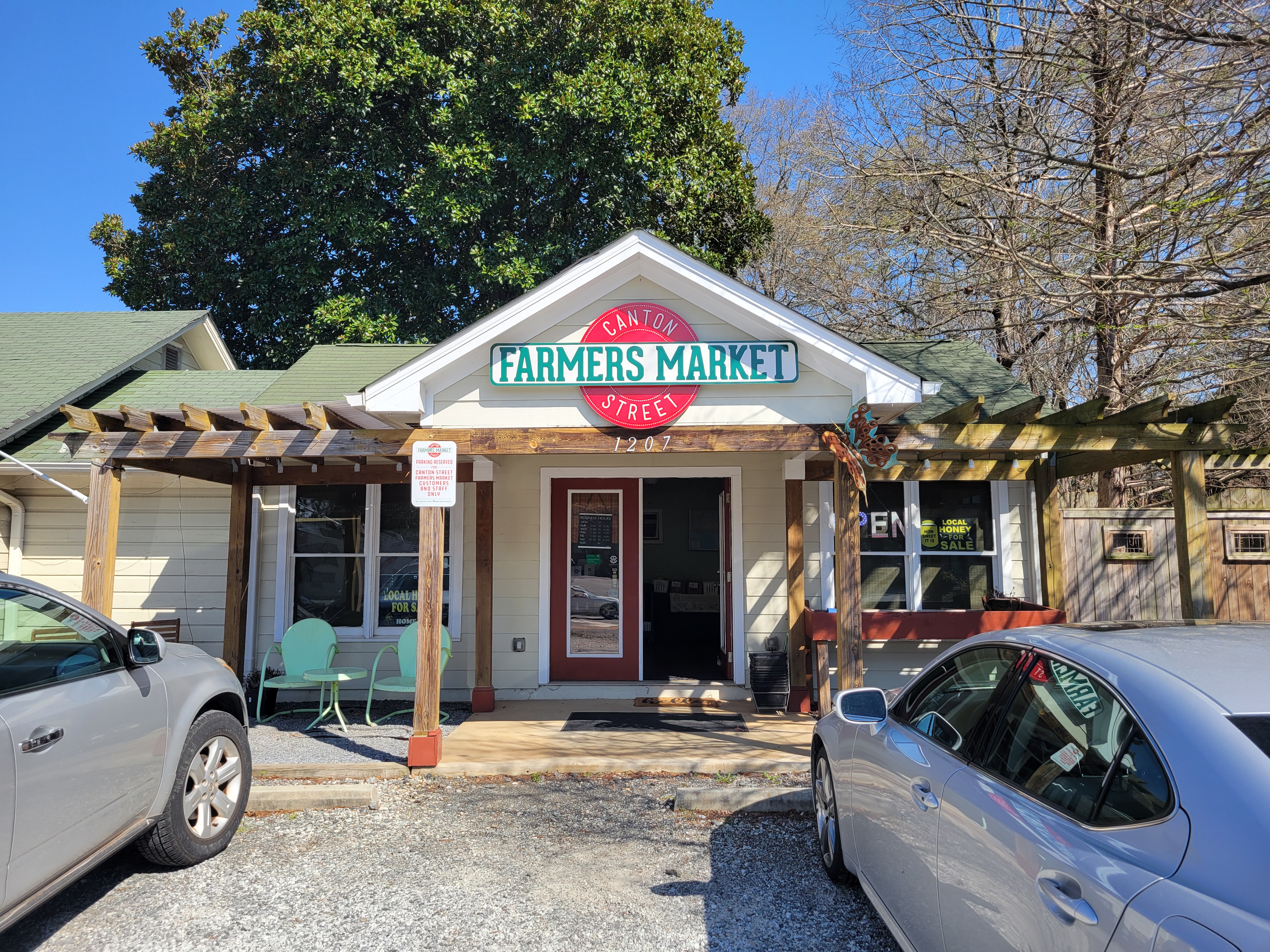 Small building with a covered porch and sign reading Canton Street Farmers Market, with parked cars and trees.