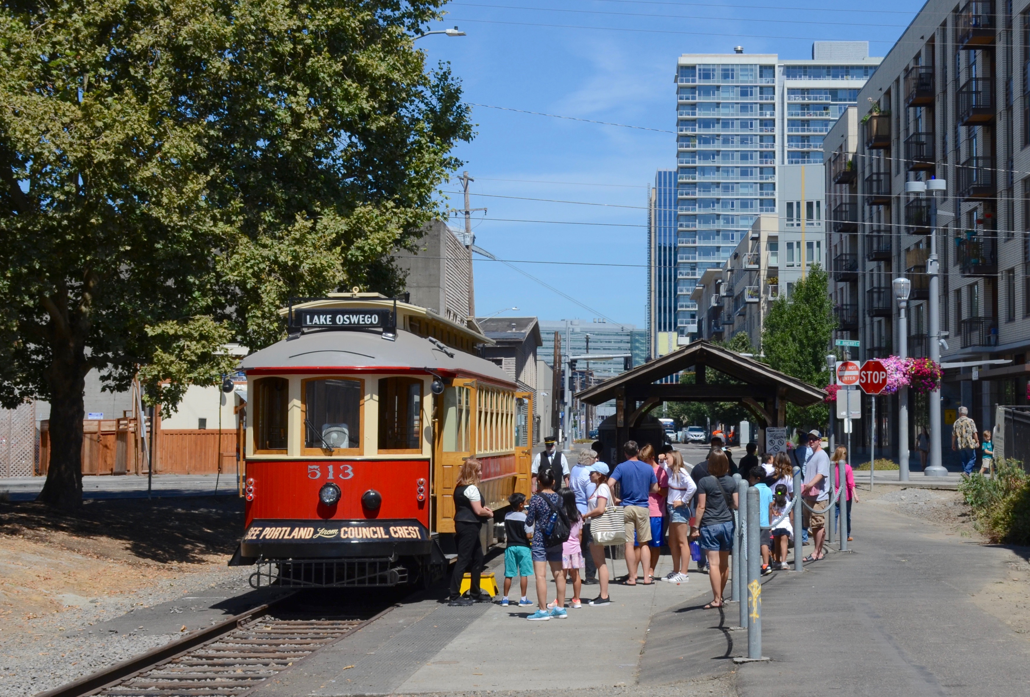 A red and cream trolley at a covered station platform with passengers boarding and city buildings behind.