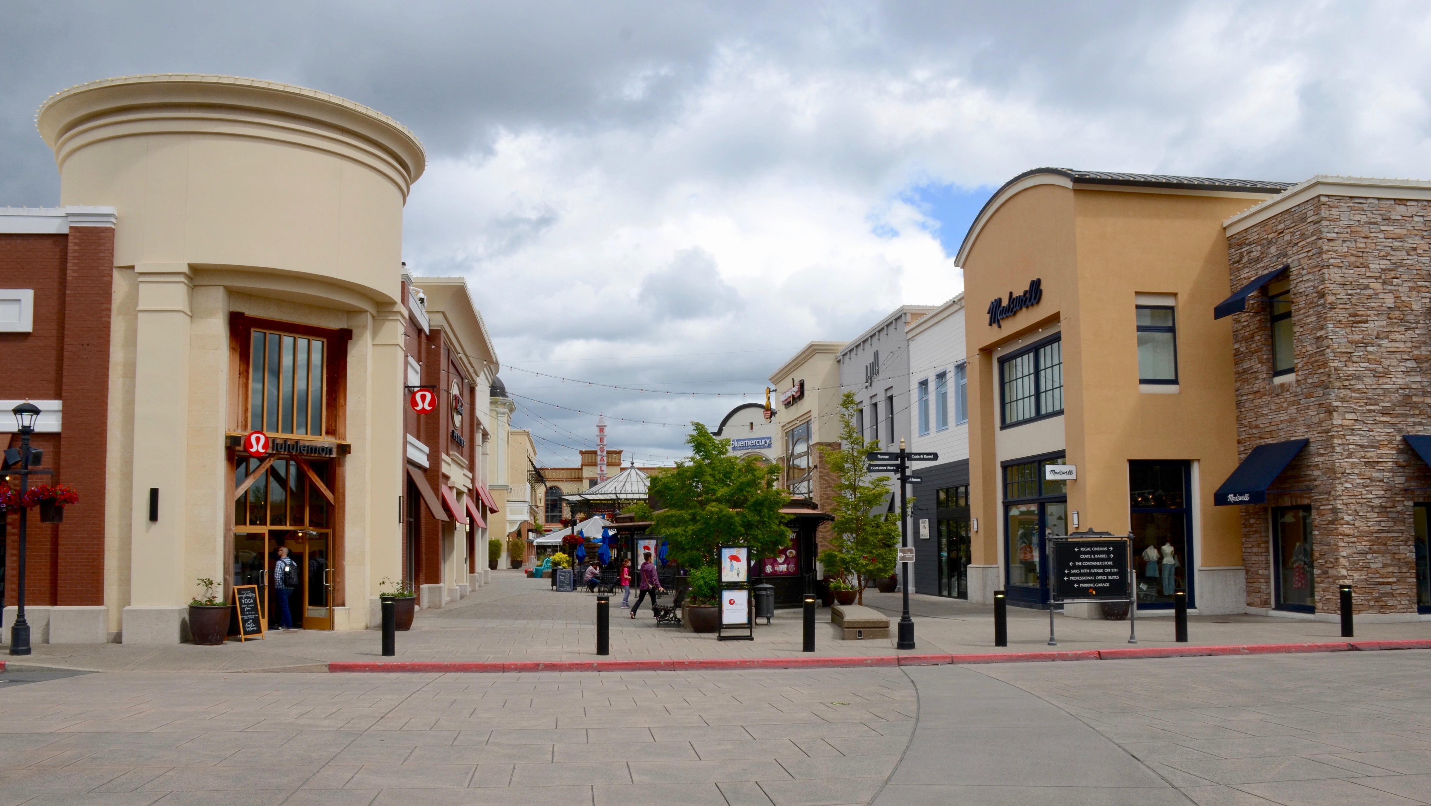 An outdoor shopping mall walkway lined with retail storefronts under a partly cloudy sky.