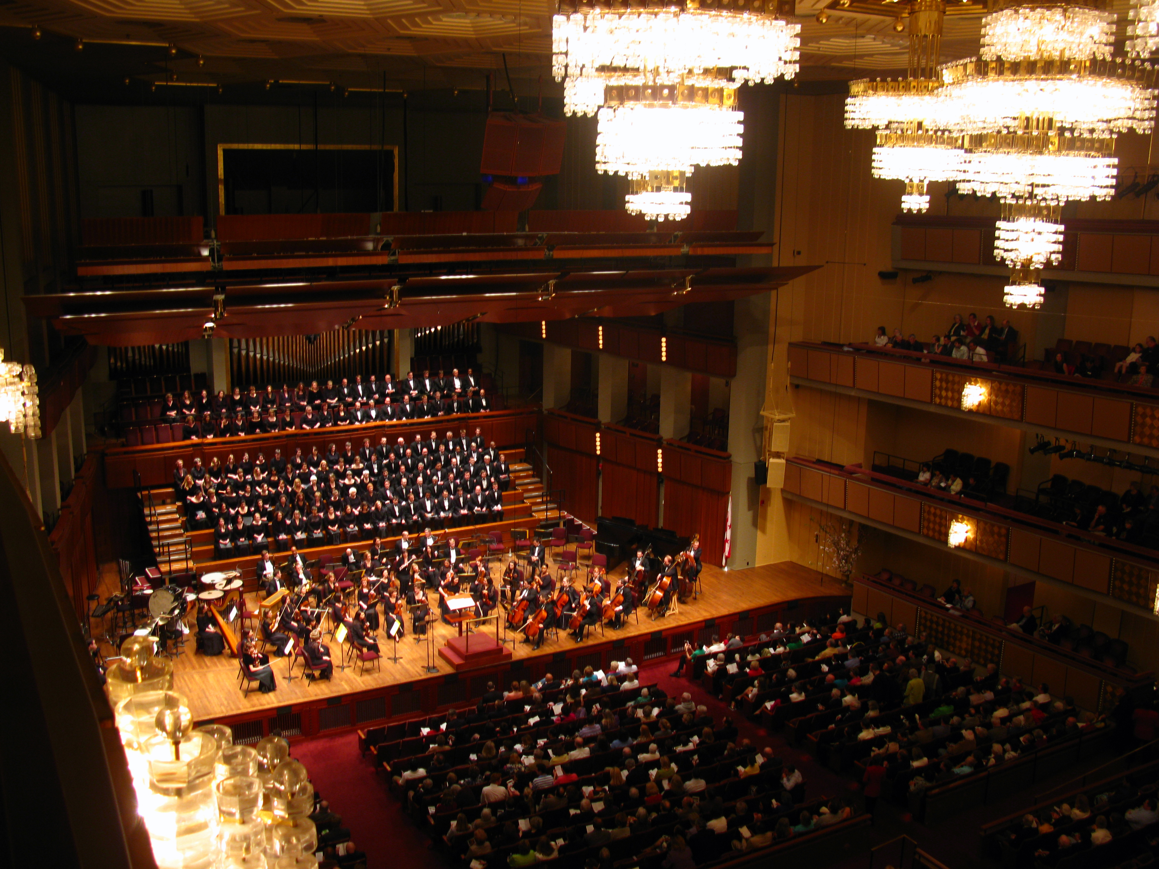 Orchestra and choir performing on stage in a large concert hall viewed from an upper balcony.