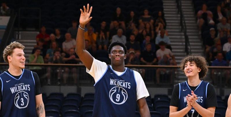 Three basketball players in Villanova jerseys stand on a court, center player waving.