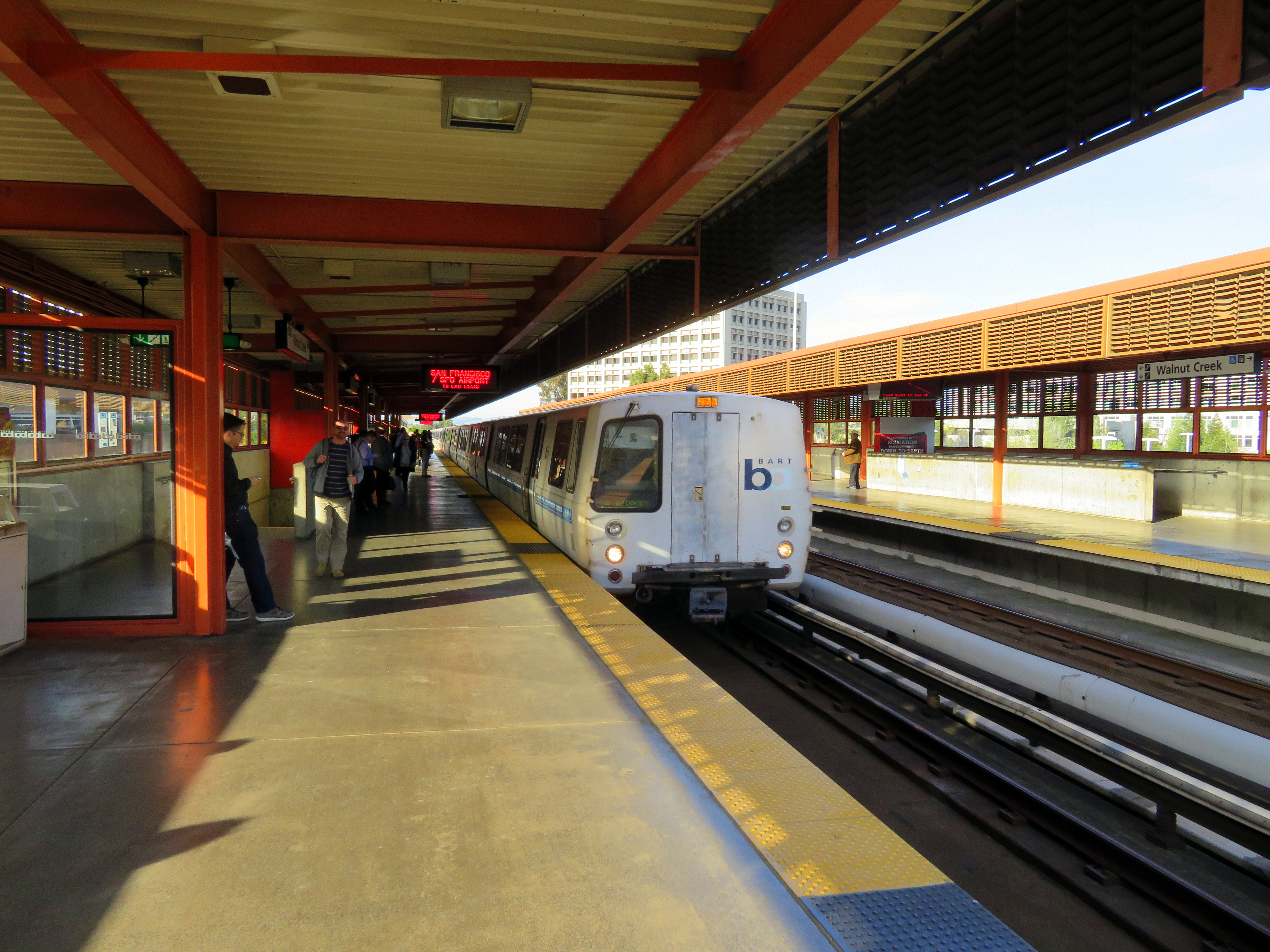 A BART train arrives at a covered outdoor station platform with waiting passengers.