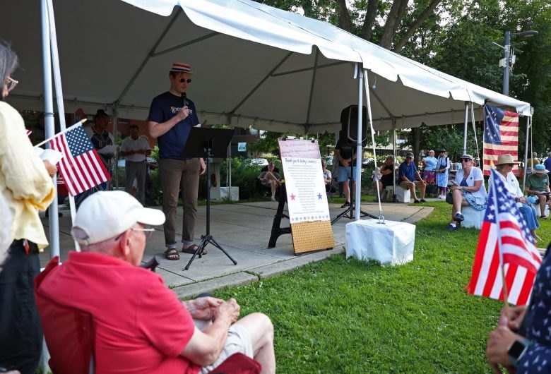 A speaker at a microphone under a white tent addresses seated audience members holding small American flags in a park.