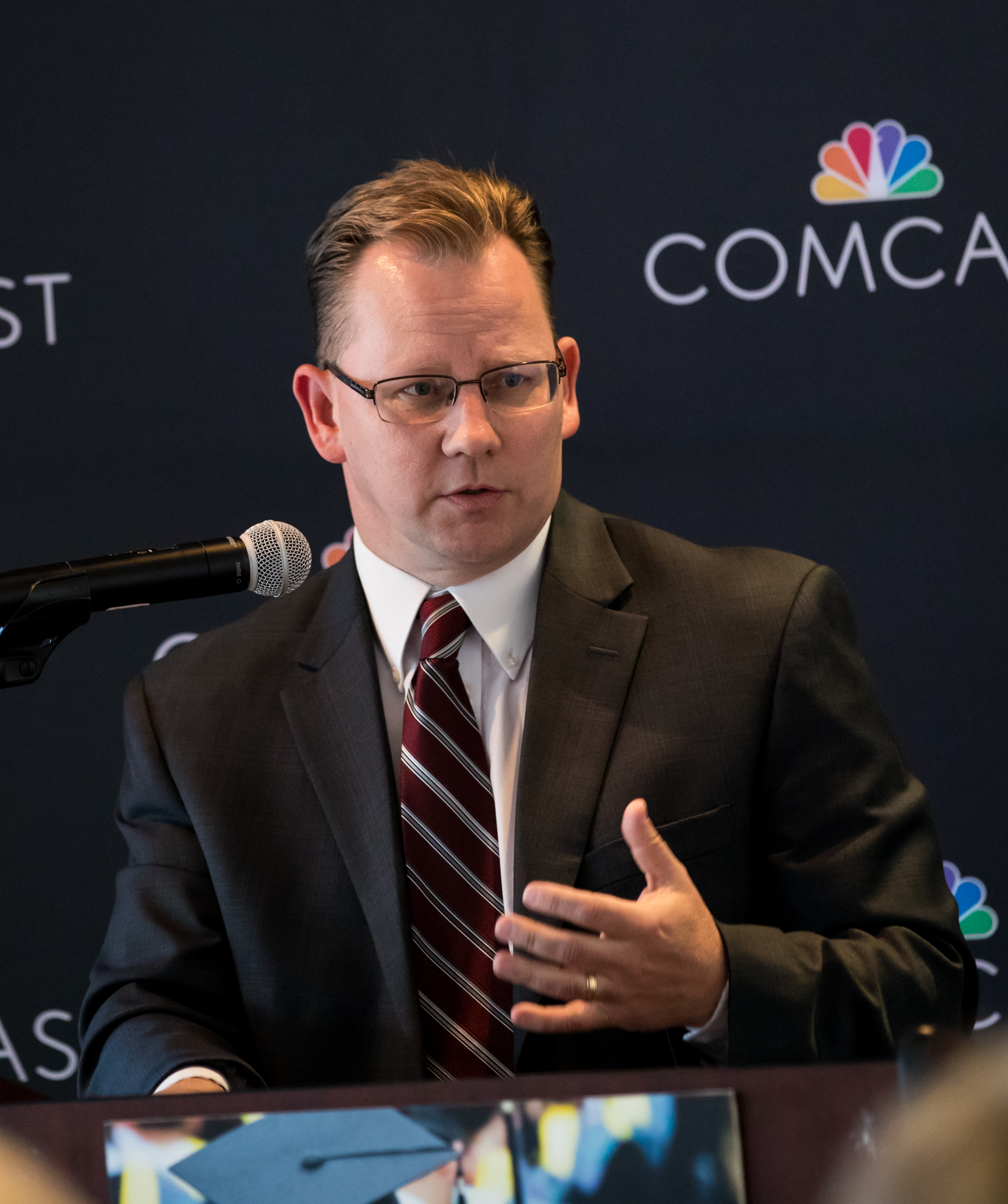 A man in a dark suit and striped tie speaks at a podium in front of a branded event backdrop.
