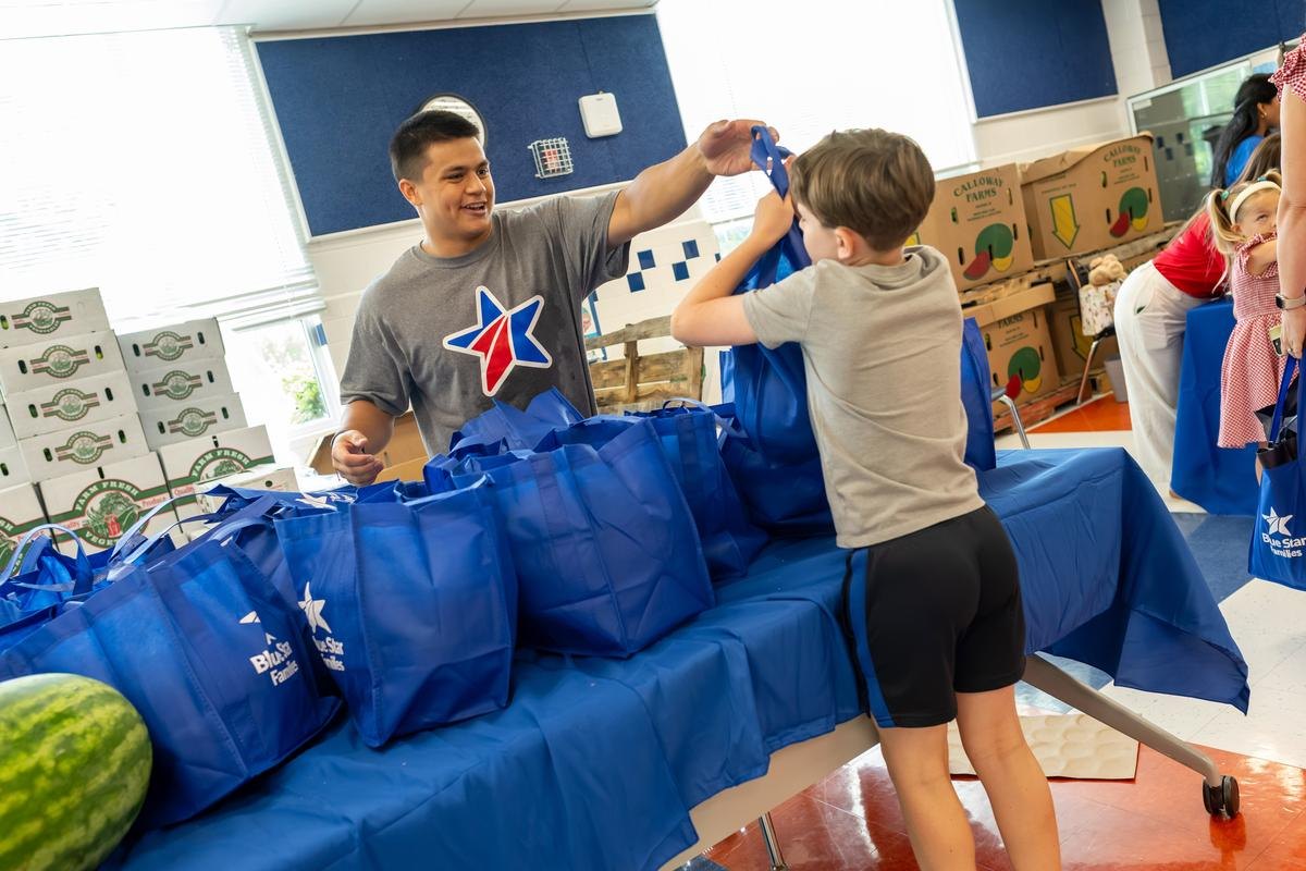 Volunteer in Blue Star Families shirt hands a blue tote bag to a child across a table.