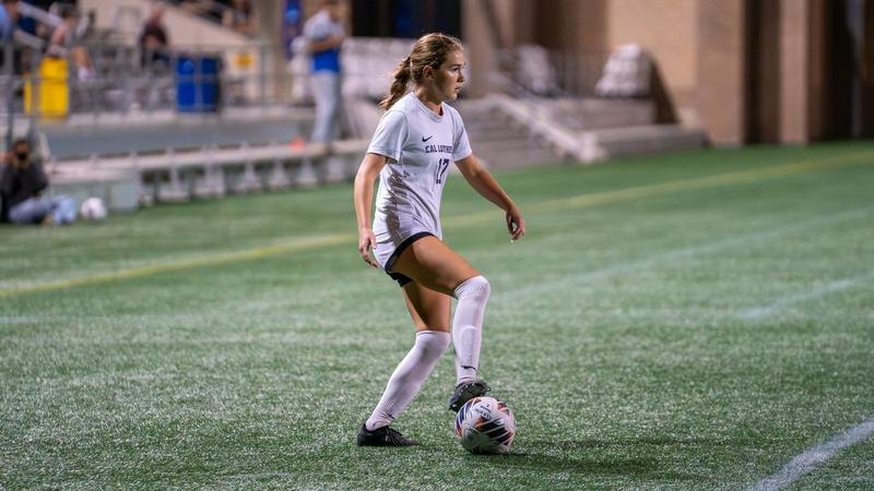 A female soccer player in a white uniform dribbles a ball on artificial turf under stadium lights at night.