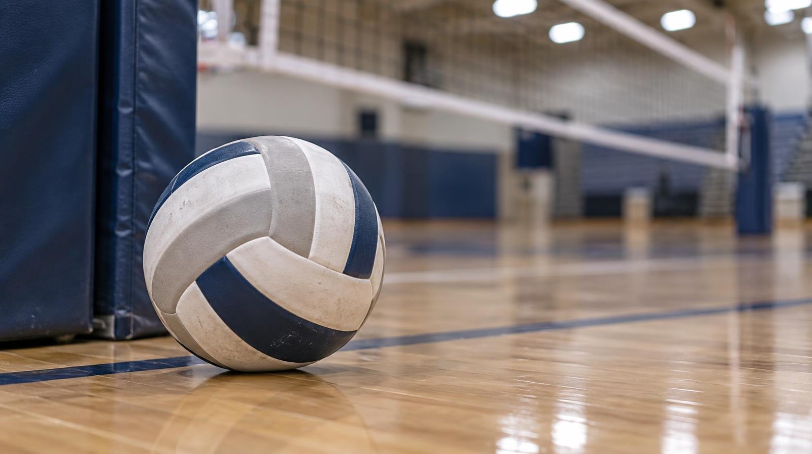 A volleyball resting on a hardwood floor near a volleyball net post.