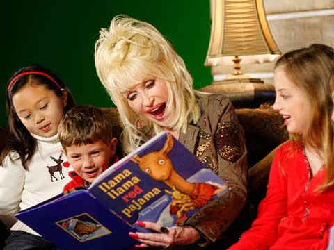 A woman with large blonde hair reads a picture book to three young children on a couch.