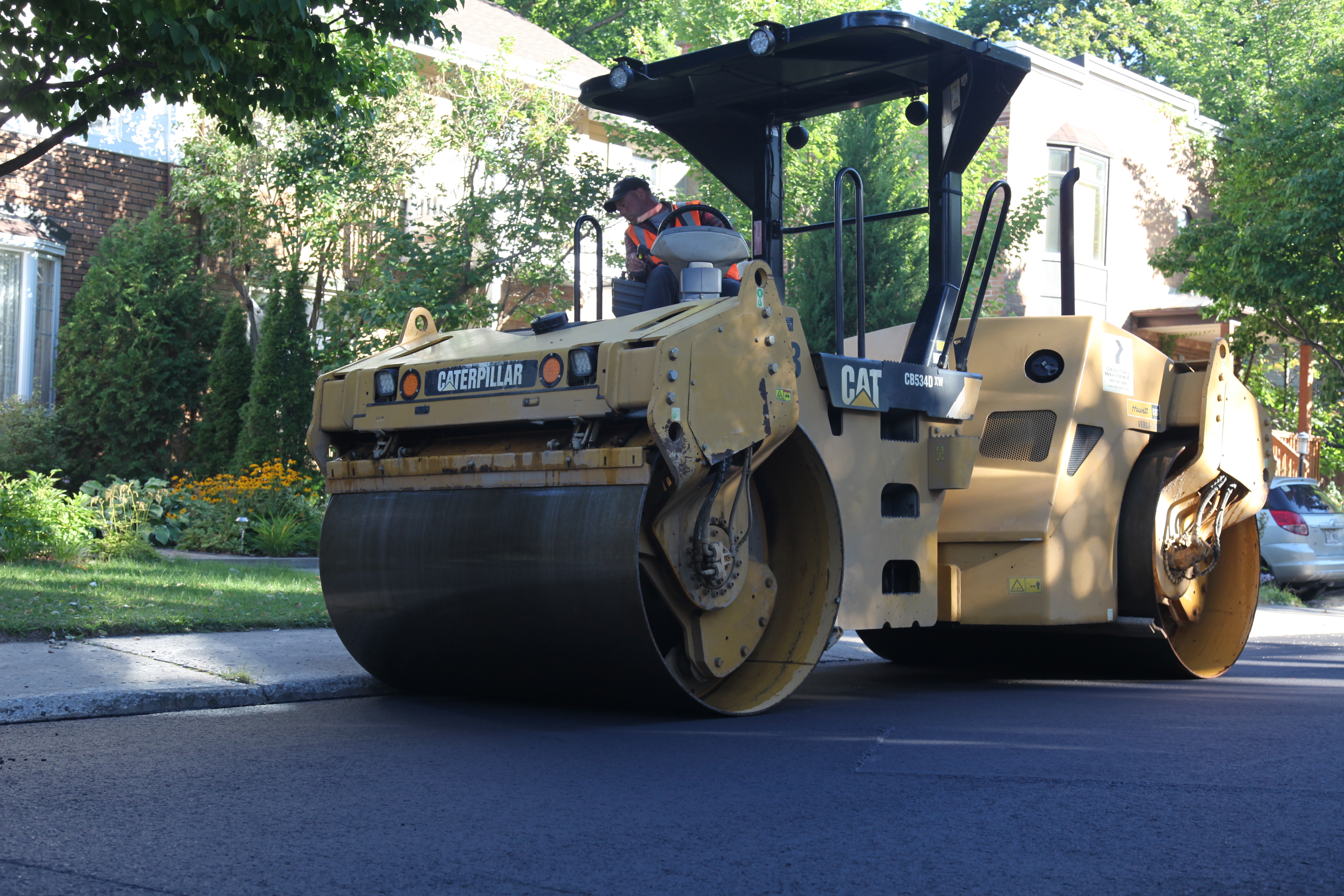 A yellow Caterpillar roller compactor compacting fresh asphalt on a residential street.