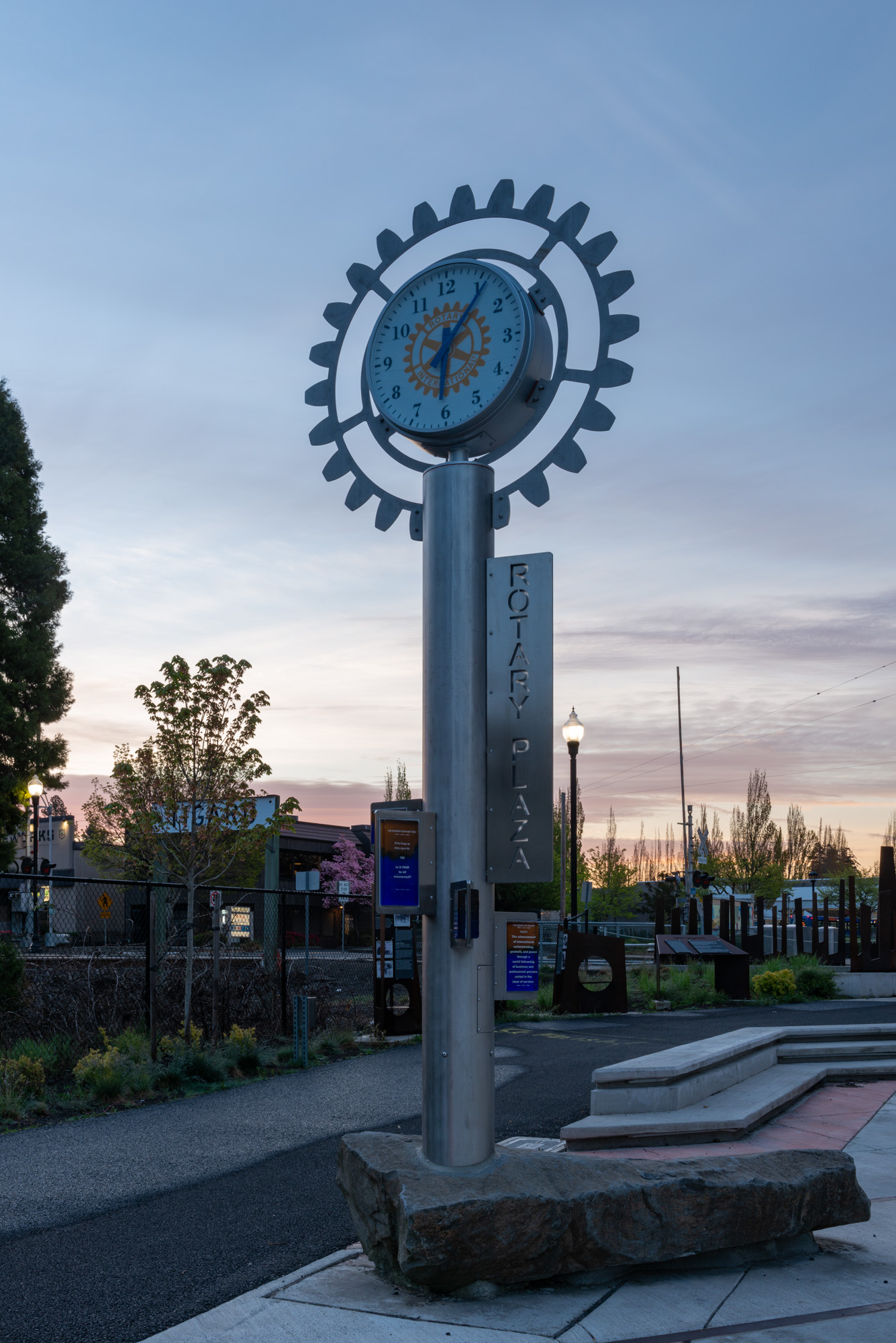 A gear-shaped clock on a metal pillar along a pedestrian path at dusk.
