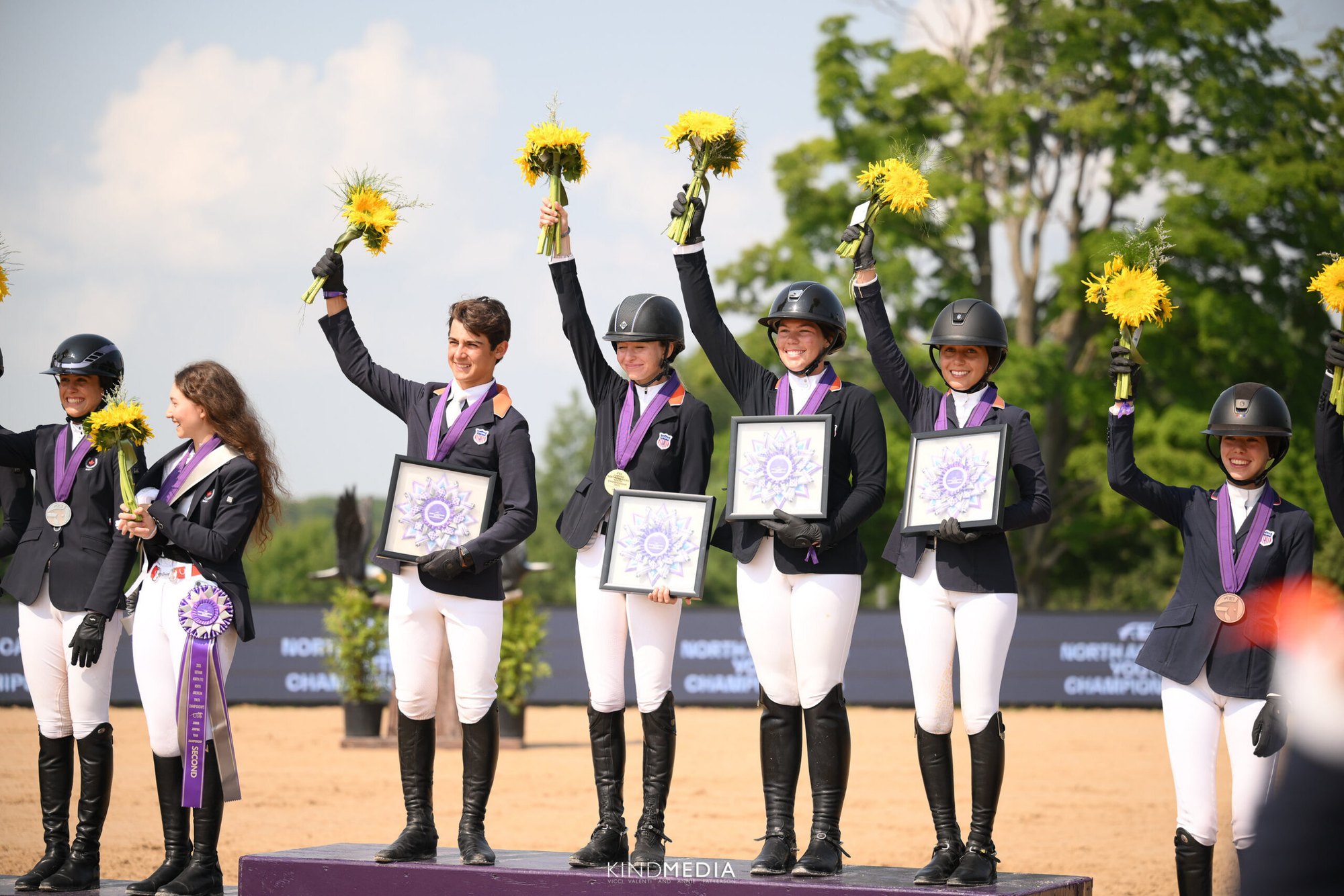 Young equestrian riders in black jackets stand on a podium holding yellow flower bouquets and medals.