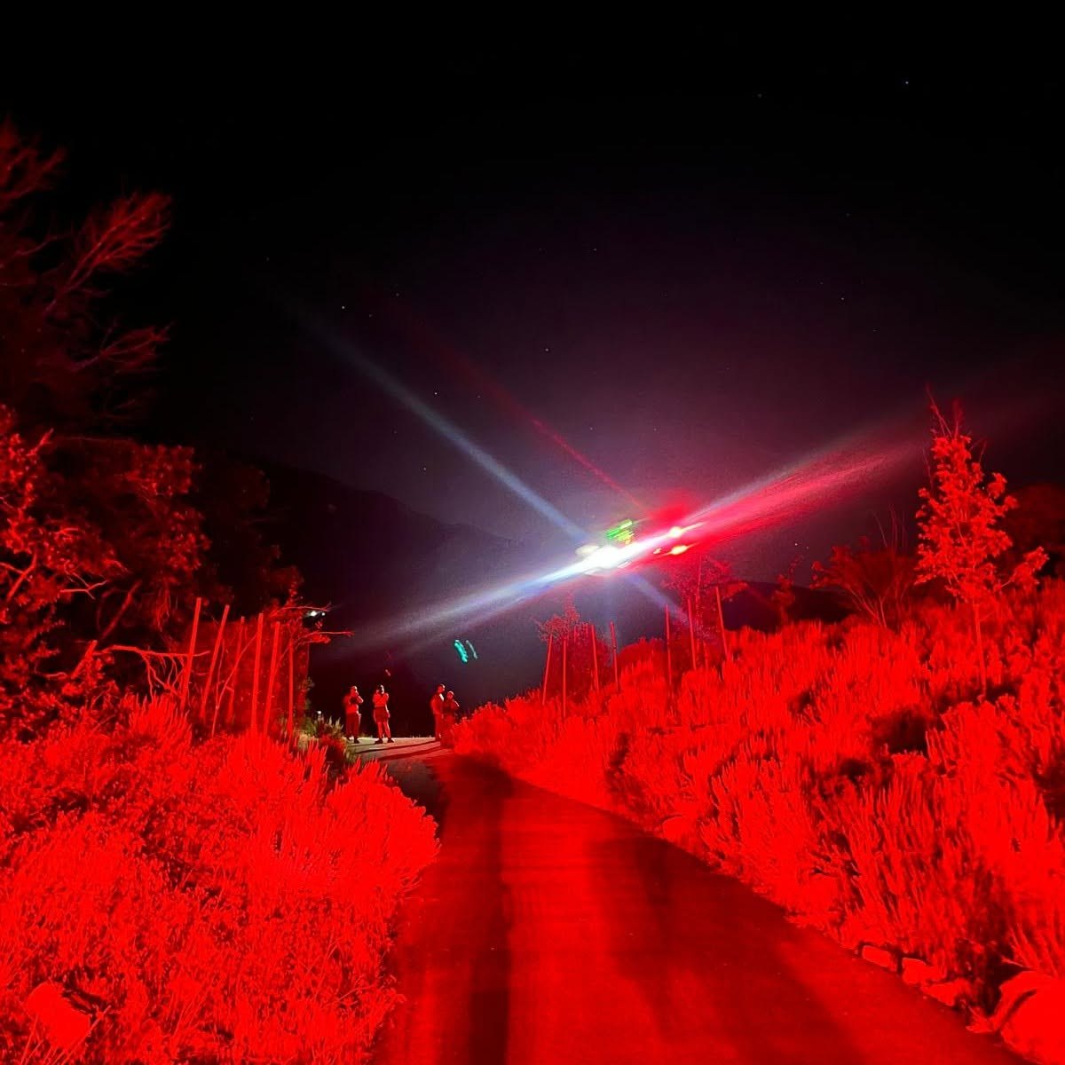 Helicopter hovering overhead with spotlight illuminating rescue personnel on a dark trail at night.