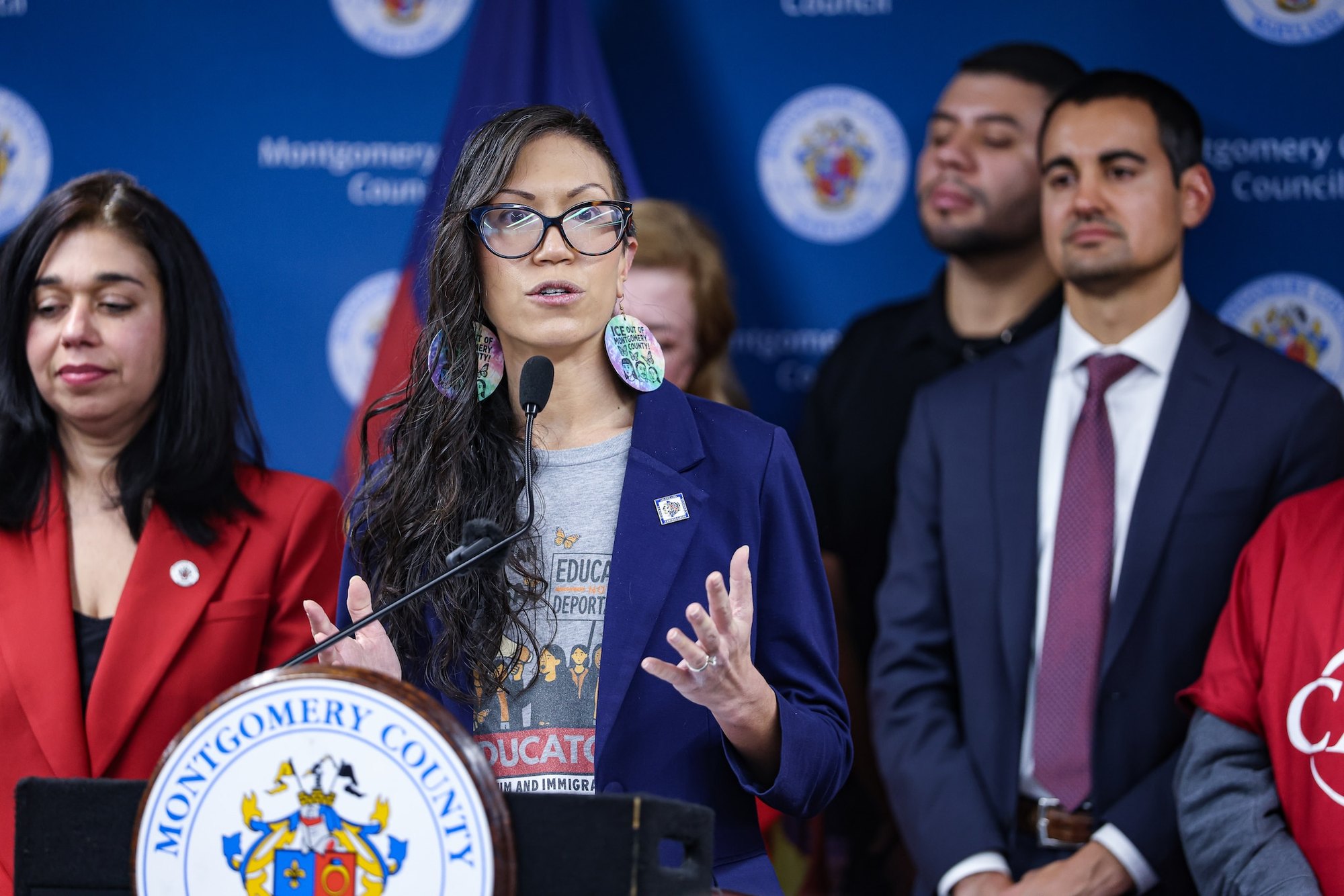 A woman in a blue blazer and glasses speaks at a podium with the Montgomery County seal.
