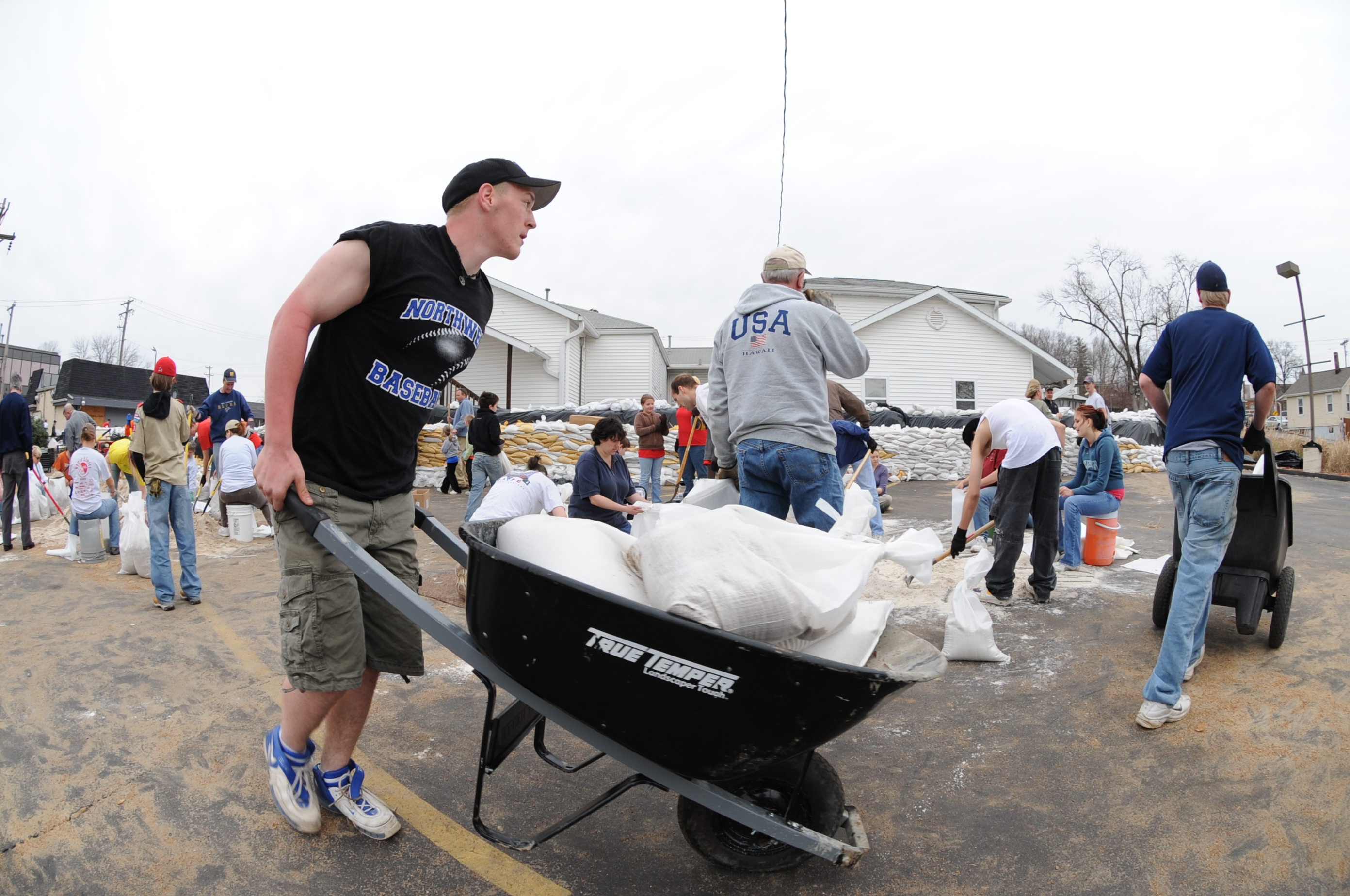 Volunteers filling sandbags and loading a wheelbarrow in a parking lot.