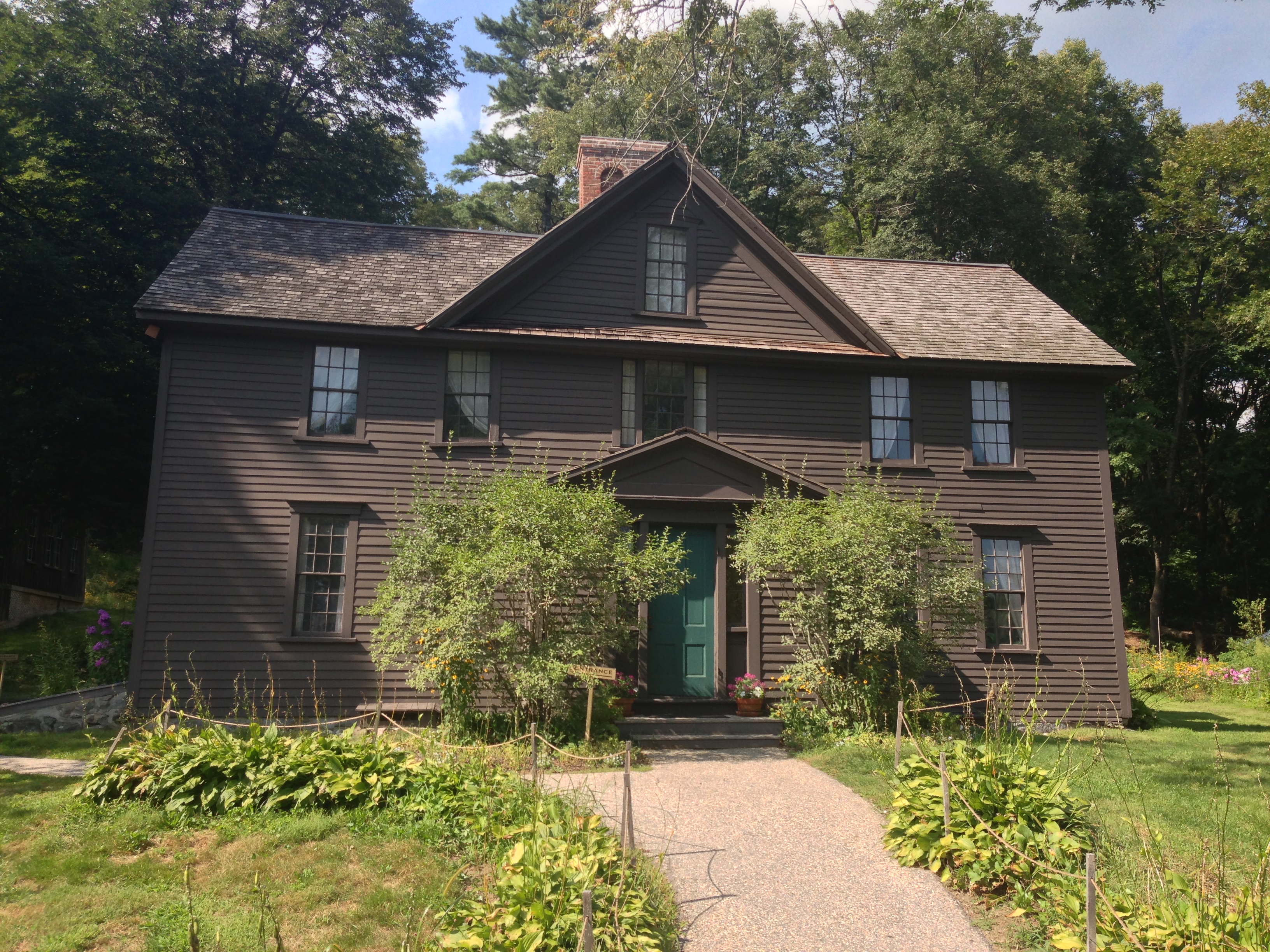 Brown two-story colonial house with teal front door and lush garden surroundings.