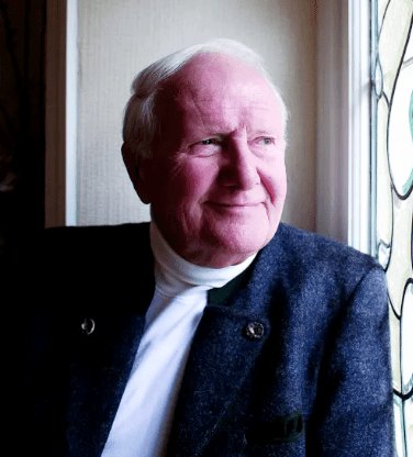 Elderly man with white hair in a white turtleneck and tweed jacket smiling near a stained glass window.