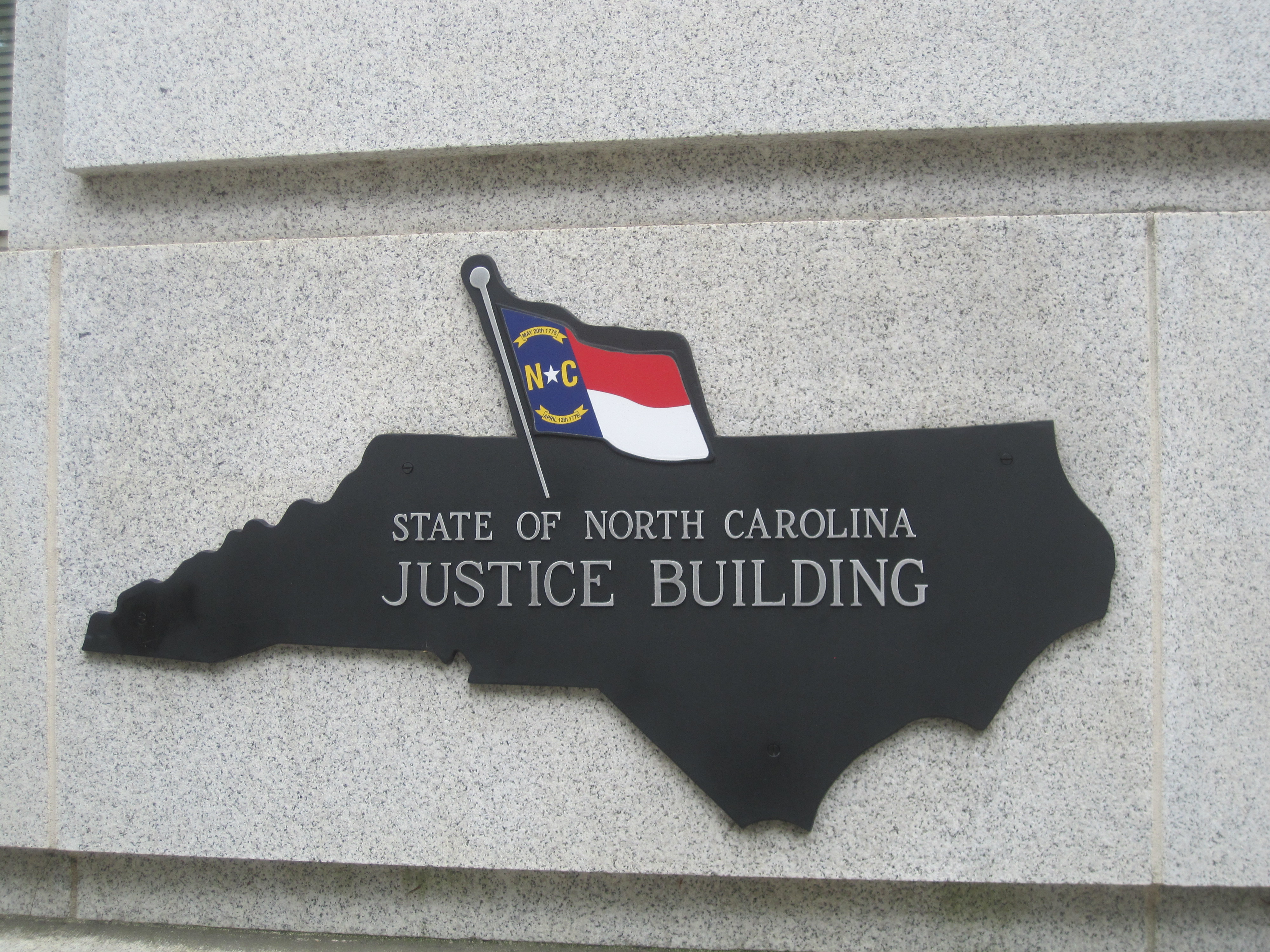 Metal sign shaped like North Carolina mounted on a granite wall outside the Justice Building.