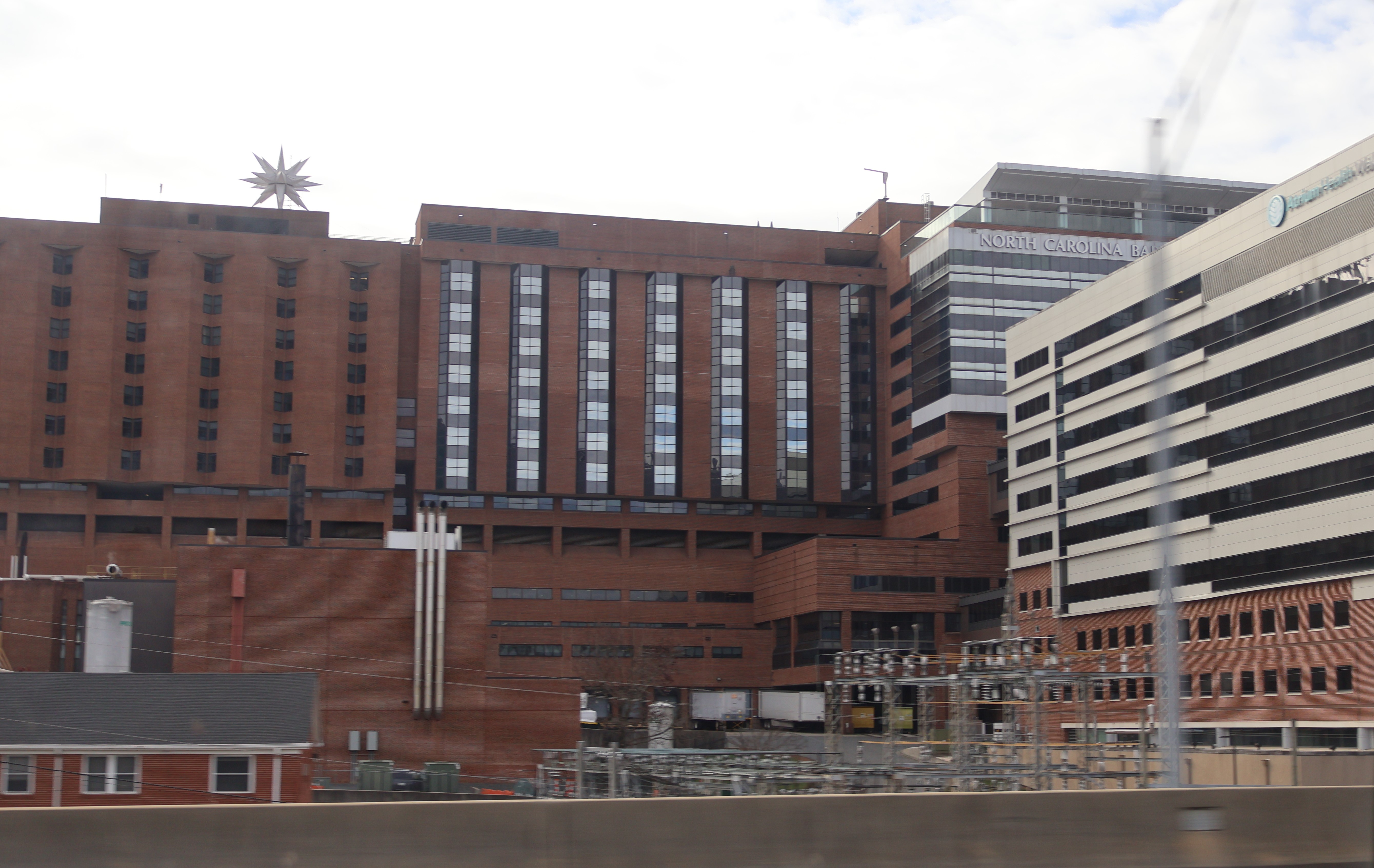 Large multi-story brick hospital complex with a cross on the facade and a newer glass addition.