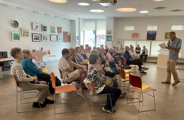 A man stands reading from papers to a seated audience of older adults in a colorful community room.