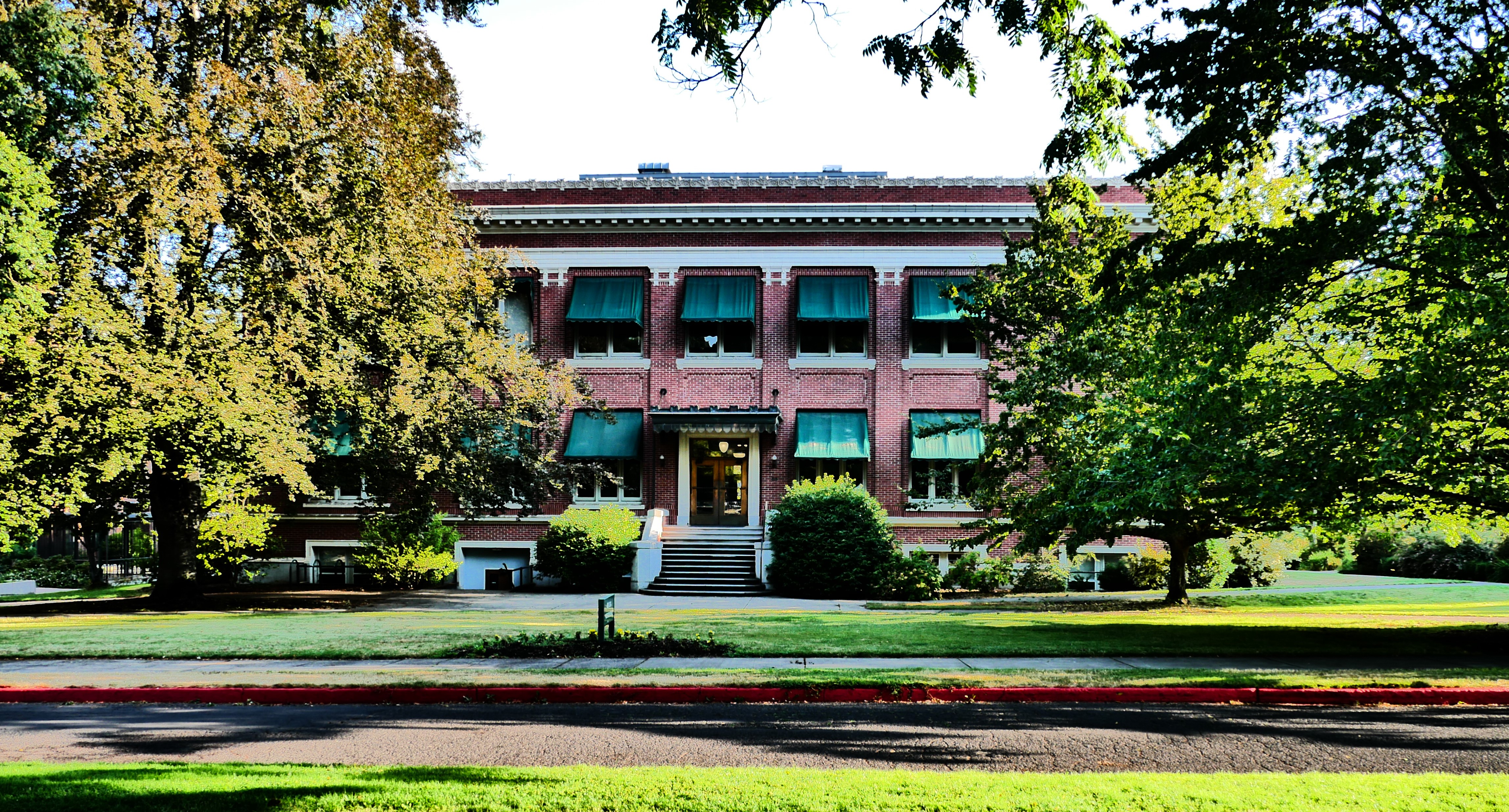 Red brick two-story academic building with green awnings surrounded by mature trees on a university campus.