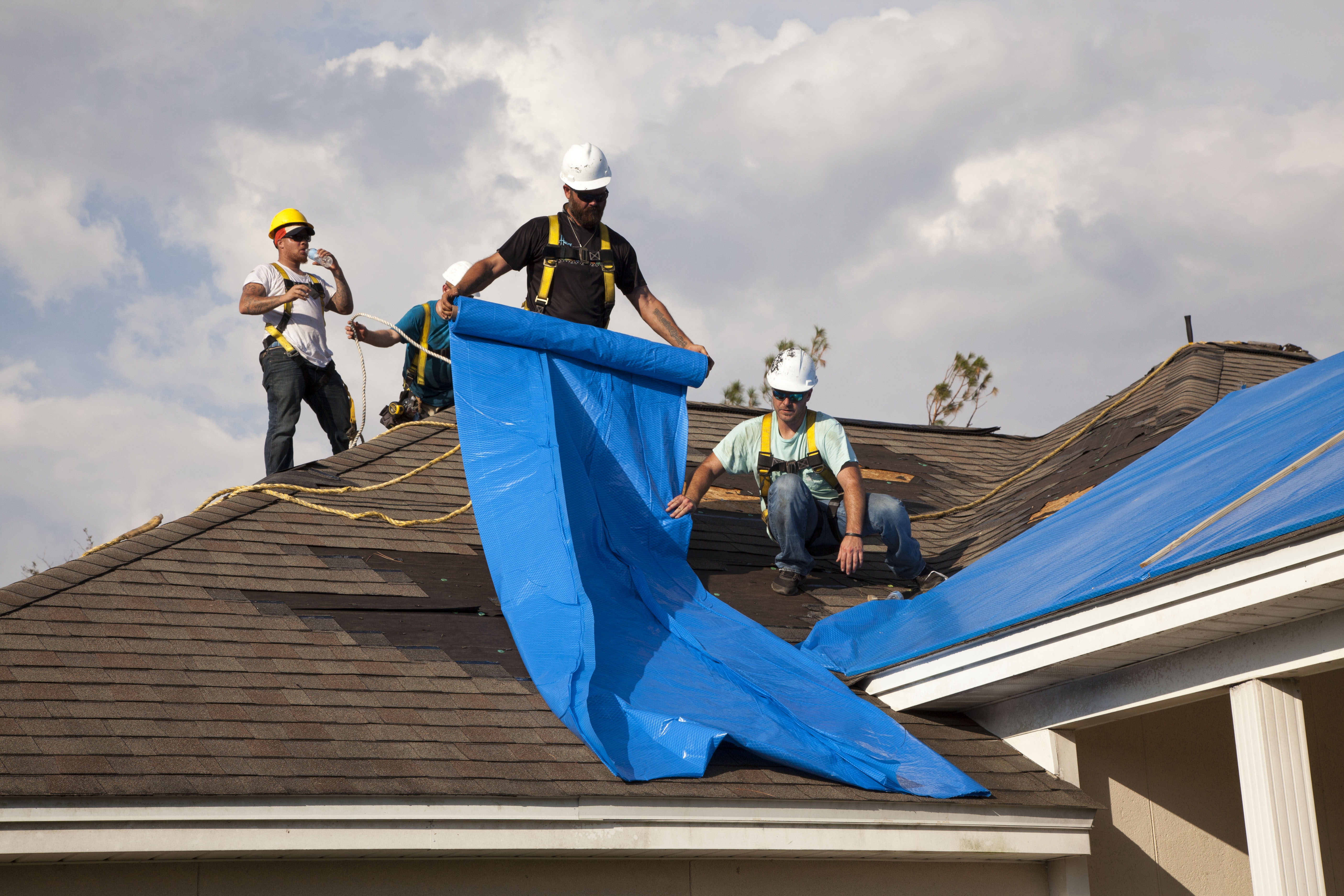 Three workers in hard hats unroll a blue tarp over a storm-damaged residential roof.