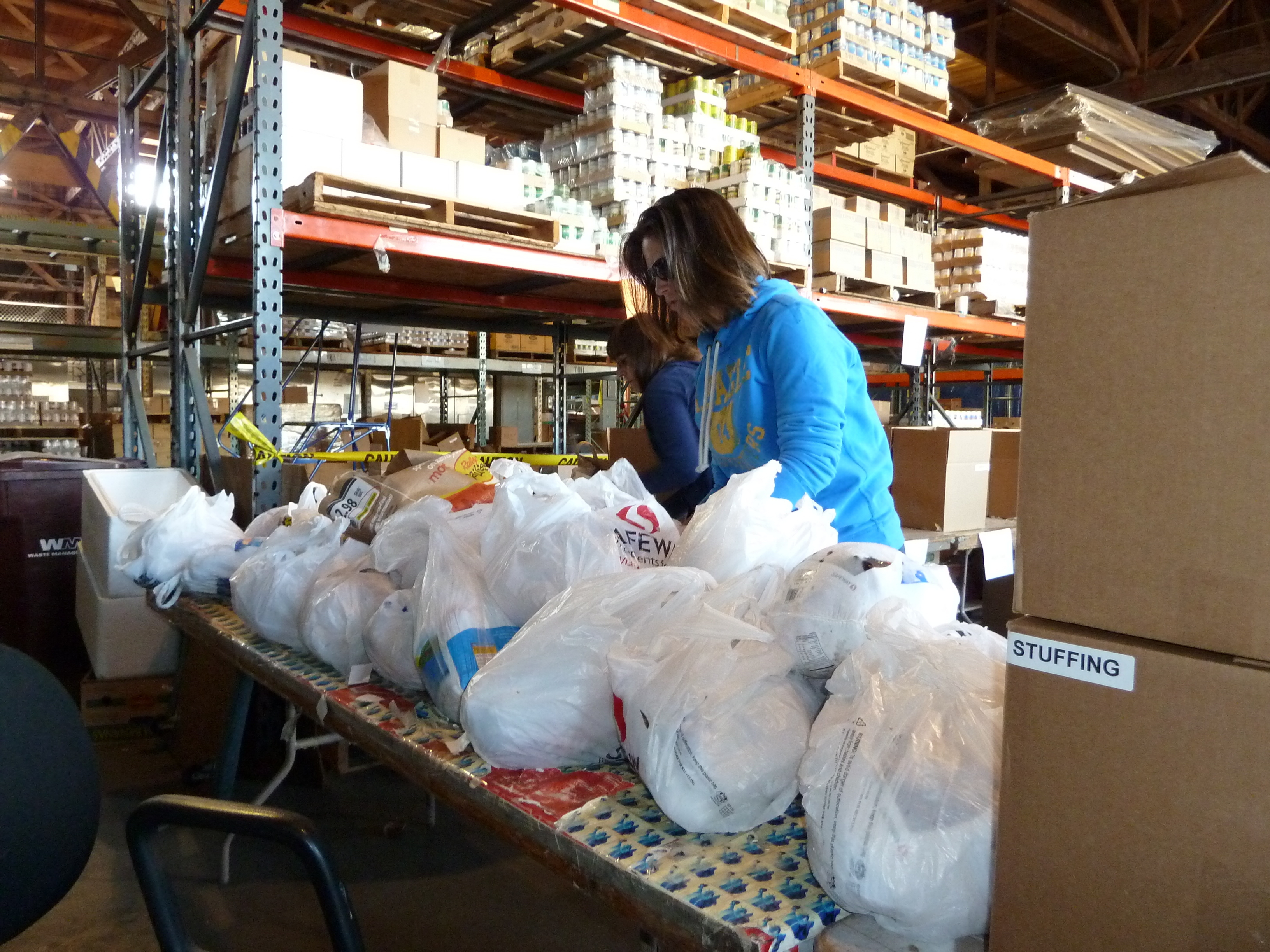 A volunteer in a blue hoodie sorts bags of donated food on a warehouse table.
