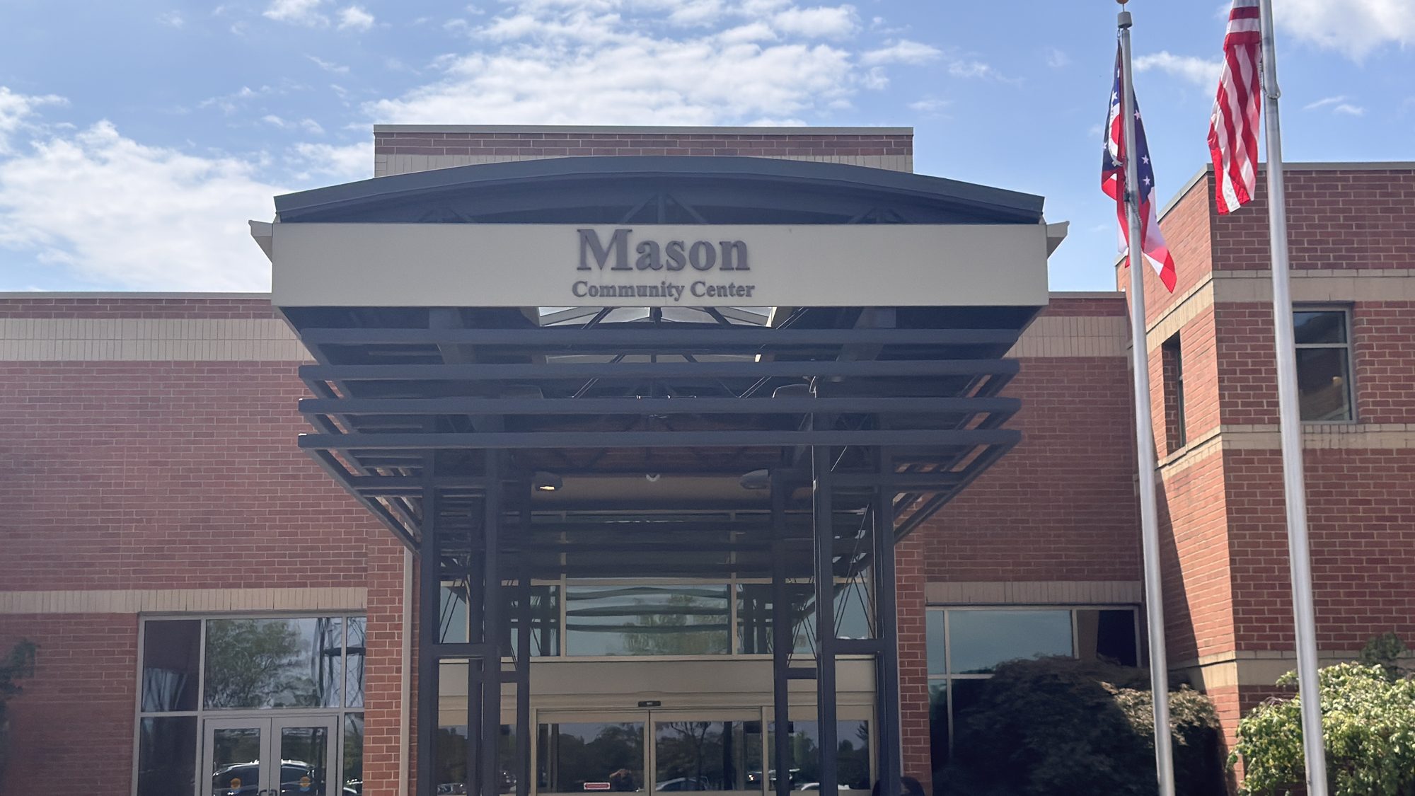 Brick community center building with covered entrance and two flagpoles under a partly cloudy sky.