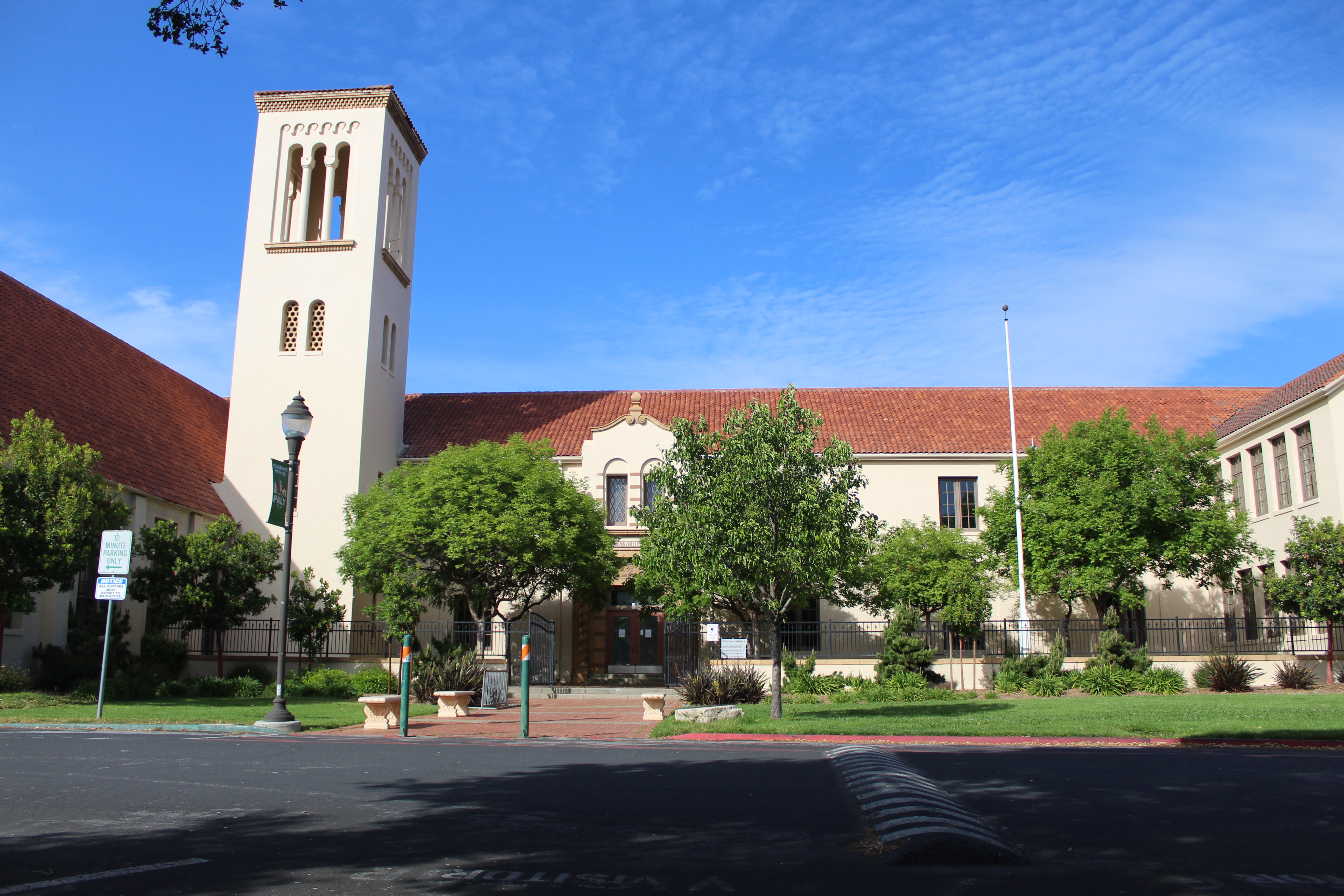 Beige stucco school building with a bell tower and red tile roof, trees in front.