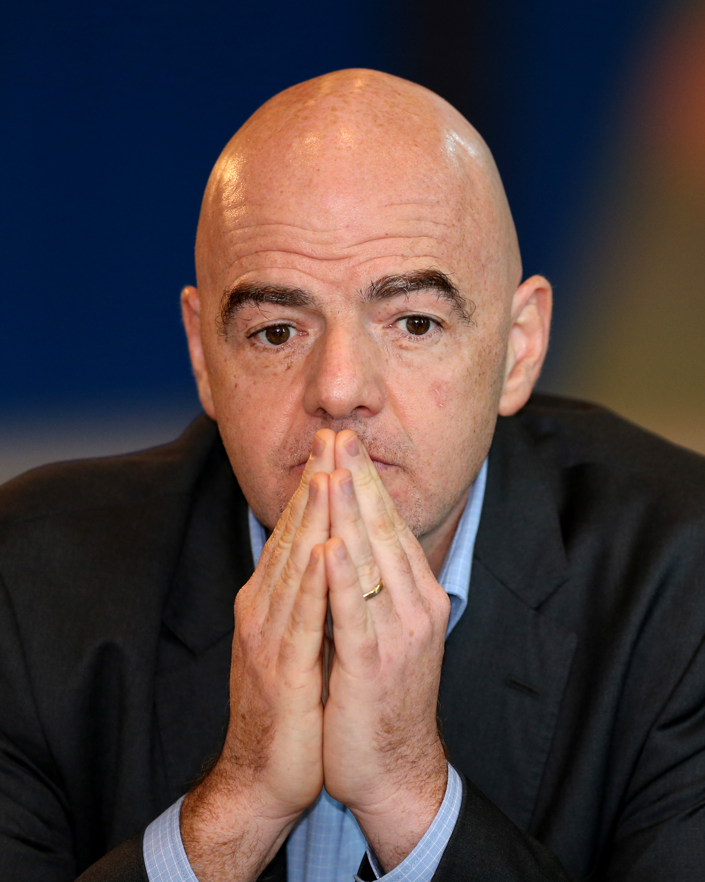 Bald man in dark suit with hands pressed together near his mouth at a press conference.