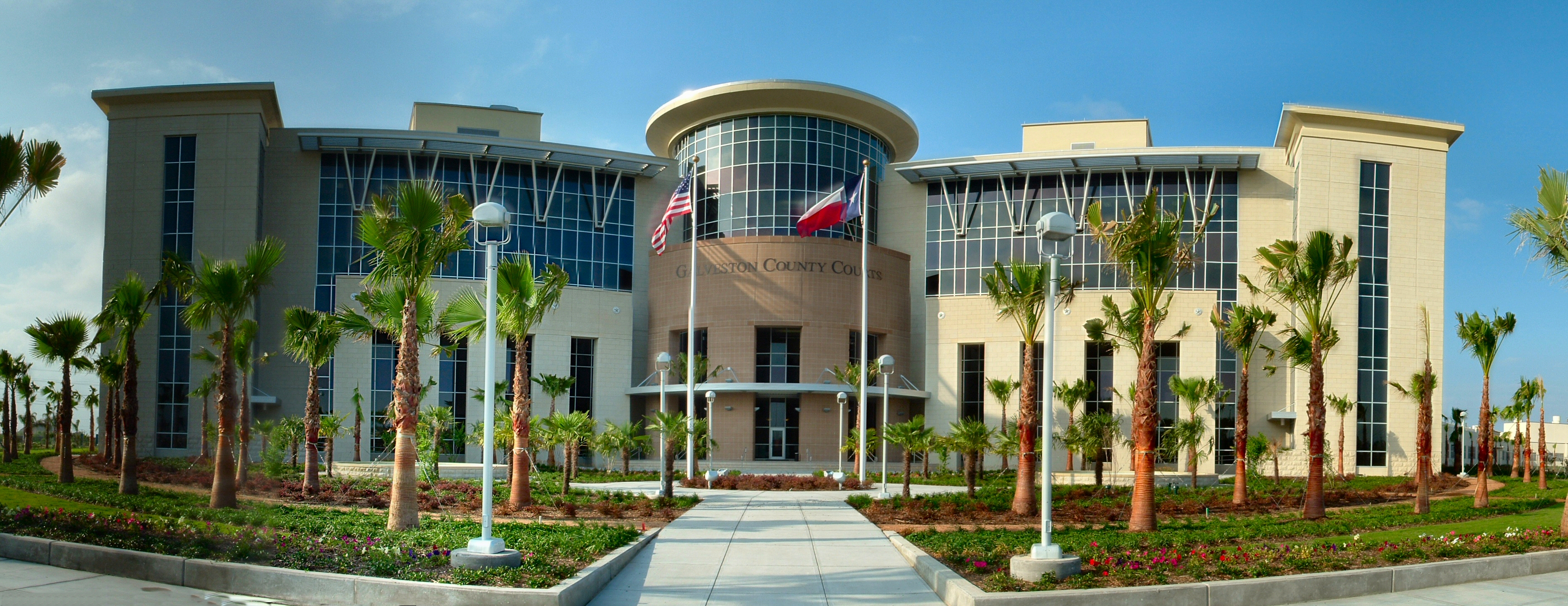 Modern government building with curved glass entrance flanked by palm trees and two flags.