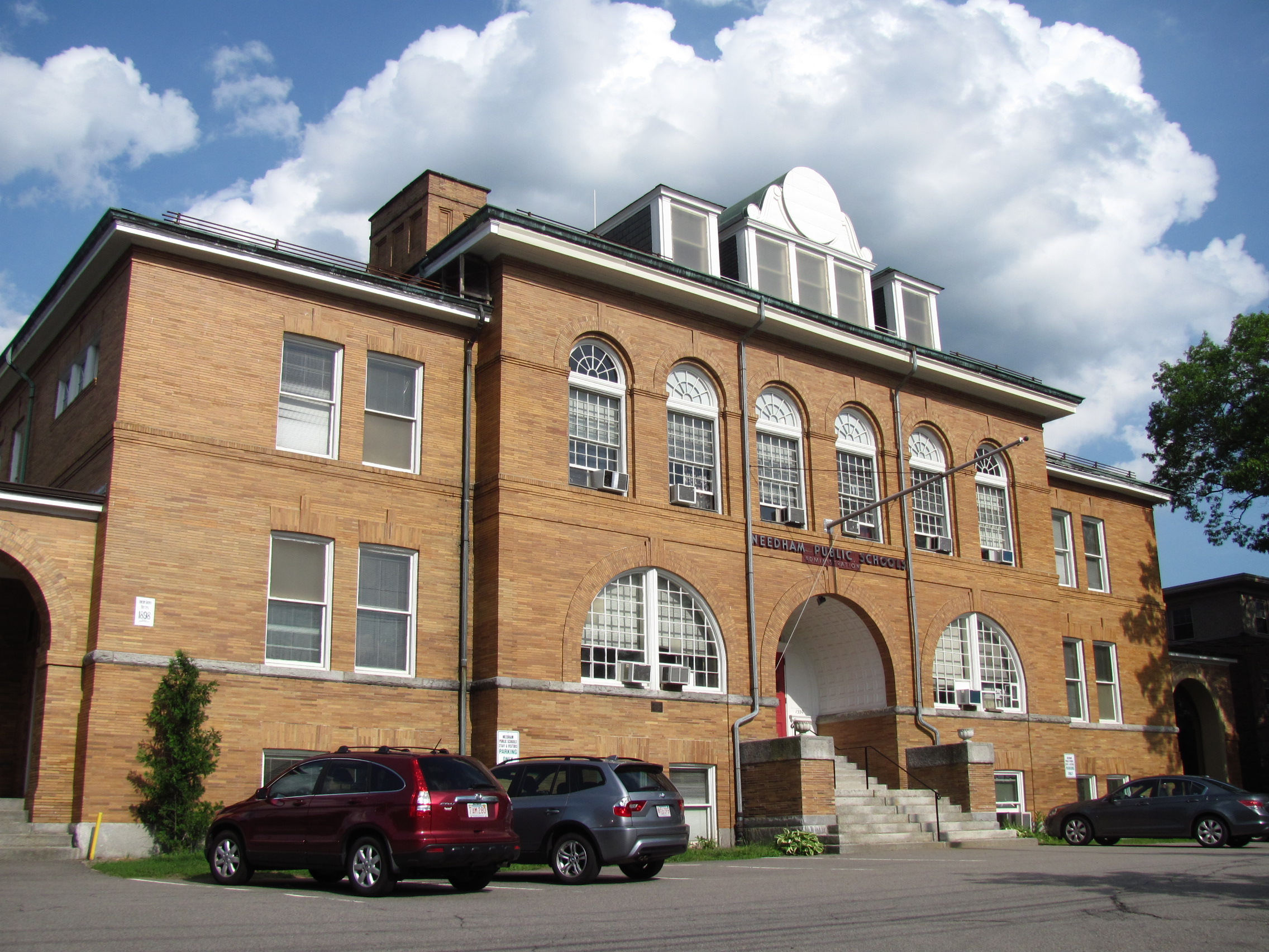 Two-story tan brick building with arched windows and central entrance steps, flanked by parked vehicles.
