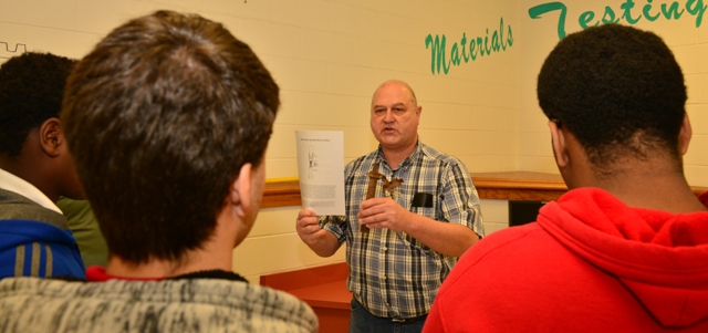 An adult presenter speaks to a small group of students gathered around him in a classroom.